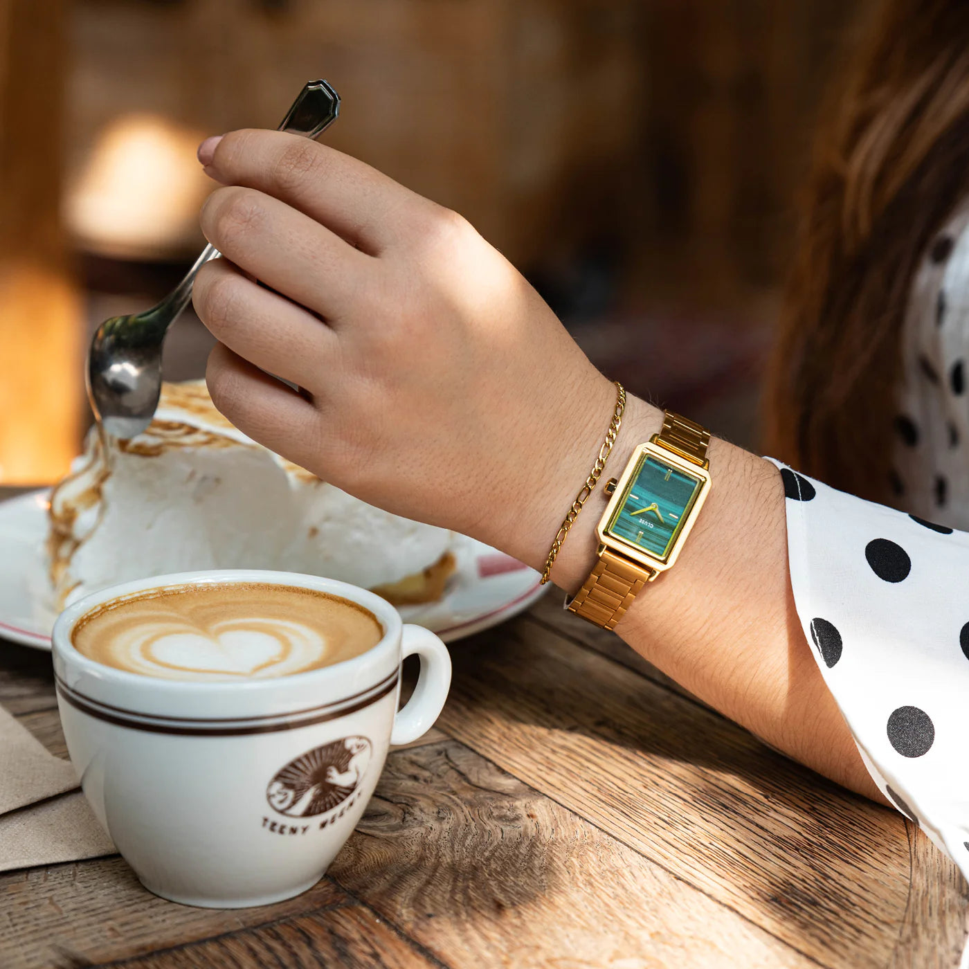 Frau trägt die Cluse "Fluette" Stahl-Uhr mit grünem Malachit-Druck-Zifferblatt mit goldfarbenem Gehäuse und kombiniert es mit einem goldenen Armband während sie einen Kuchen mit Cappocino genießt