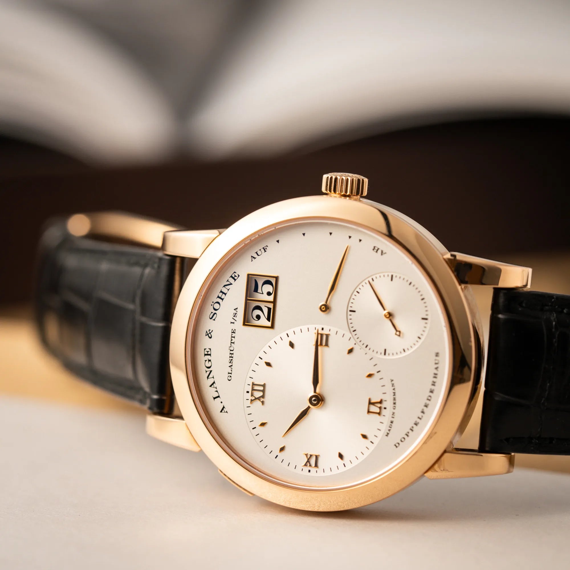 Produktfotografie mit Fokus auf dem Uhrenkopf der seitlich auf dem Gehäuse liegenden A. Lange & Söhne Lange 1 der Referenz 101.032 mit 38,5mm-Roségold-Gehäuse und dem für das Modell typische, asymmetrisch angeordnete Zifferblatt mit Großdatum