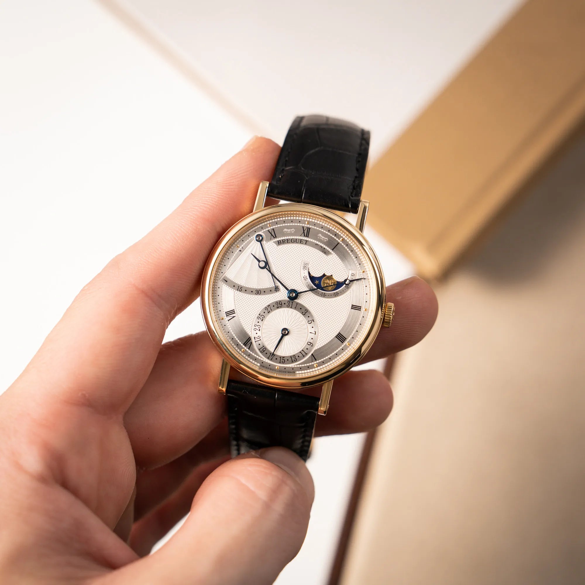 Mann hält die Breguet Classique 7137BA mit einem Gelbgoldgehäuse in klassischer Form mit einem silbernen Zifferblatt mit jeweils einem anderen handgefertigten Guilloche-Muster für jede Komplikation in seiner Hand und präsentiert die Uhr in die Kamera