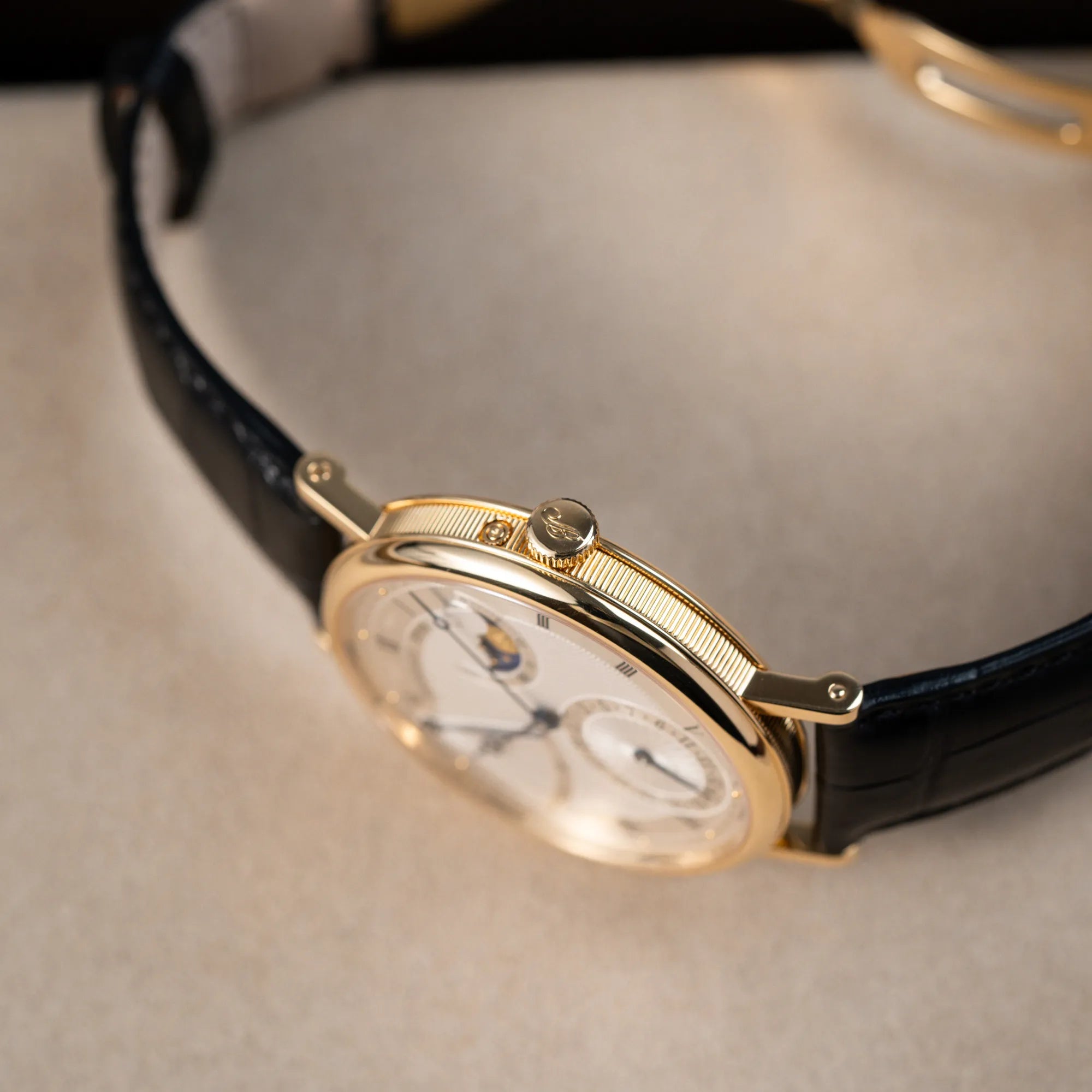 Produktfotografie der Breguet Classique 7137BA mit einem Gelbgoldgehäuse in klassischer Form mit einem silbernen Zifferblatt mit jeweils einem anderen handgefertigten Guilloche-Muster für jede Komplikation mit Fokus auf der kannelierten Gehäuseflanke und der Aufzugskrone