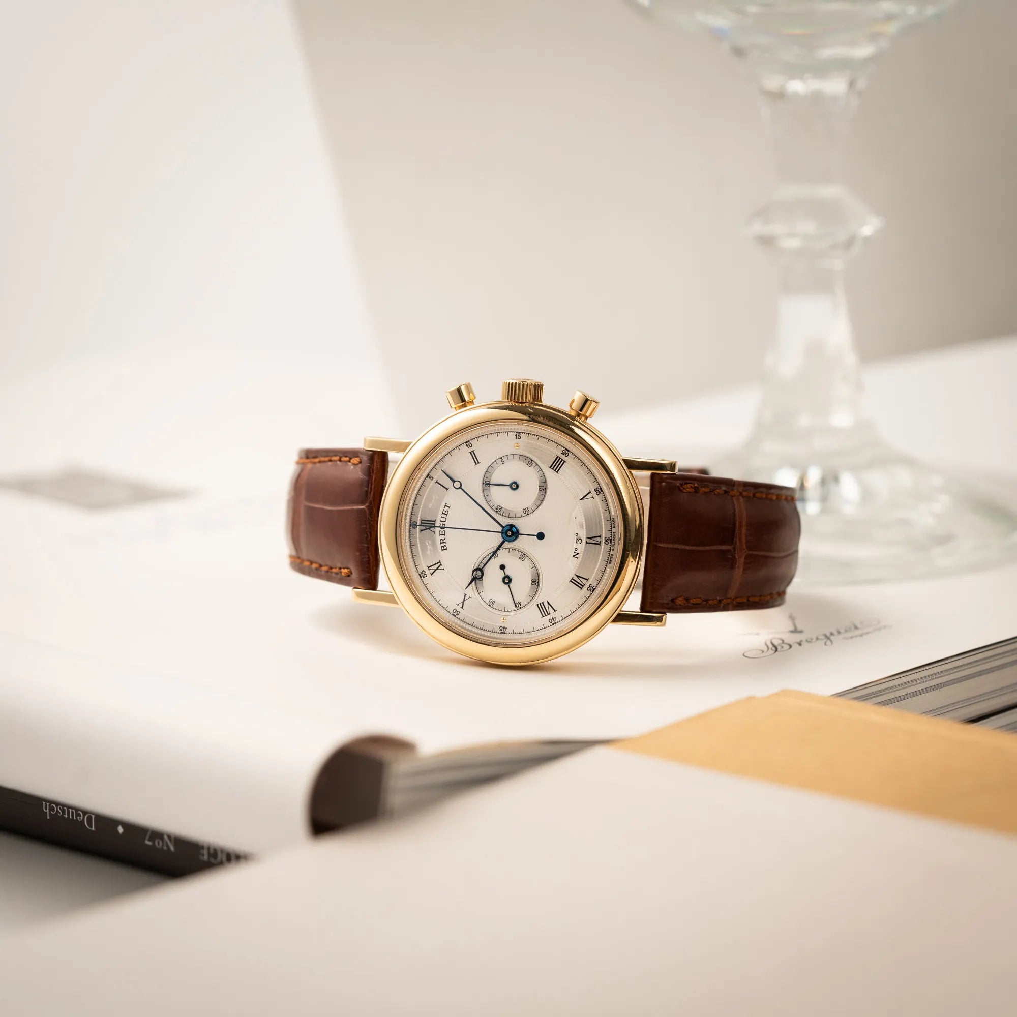 Produktfotografie der seitlich auf dem Gehäuse liegenden Breguet Classique Chronograph der Referenz 5237 in Gelbgold mit silbernem guillochiertem Zifferblatt am braunem Lederband