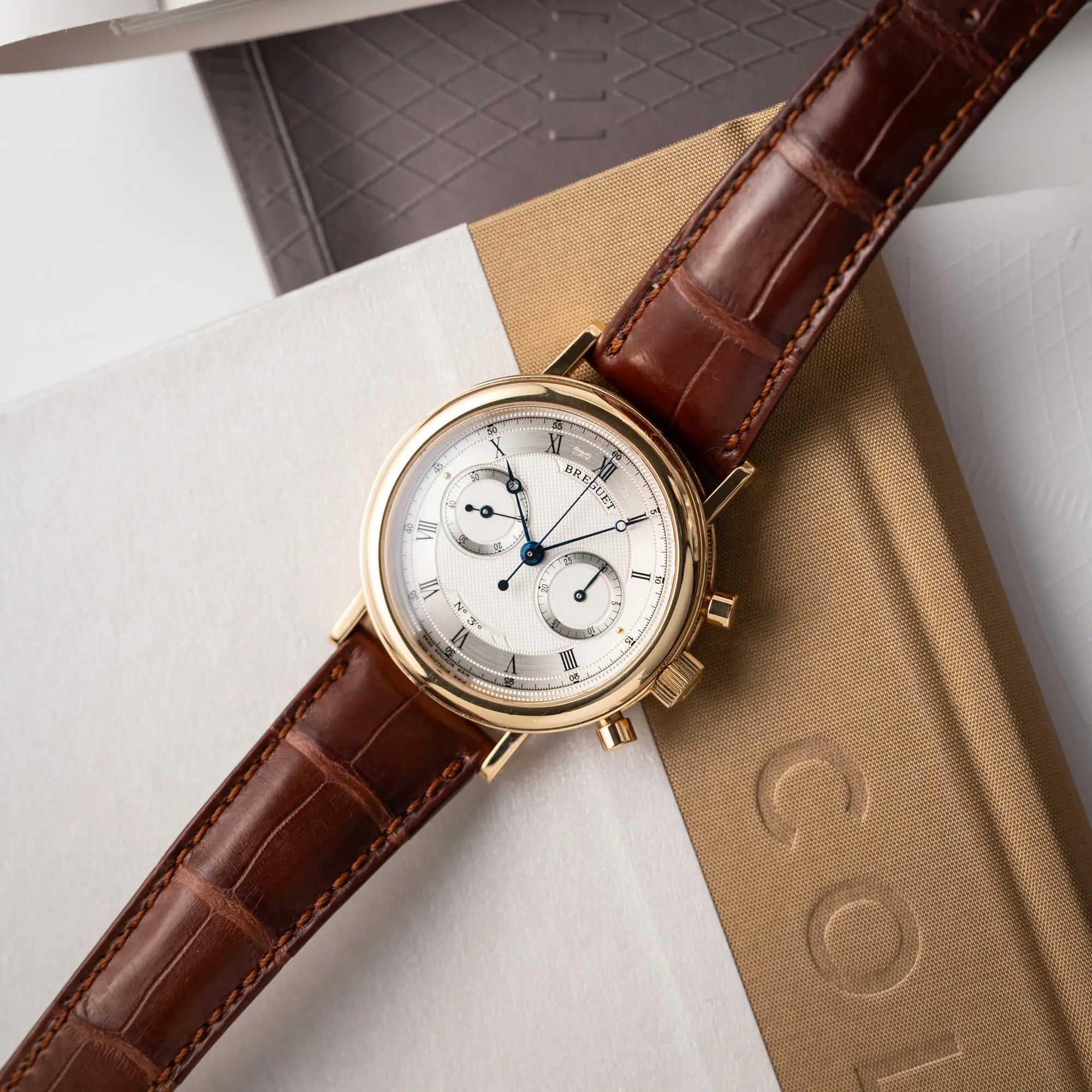 Breguet Classique Chronograph der Referenz 5237 in Gelbgold mit silbernem guillochiertem Zifferblatt am braunem Lederband liegt flach auf einem Uhrenbuch