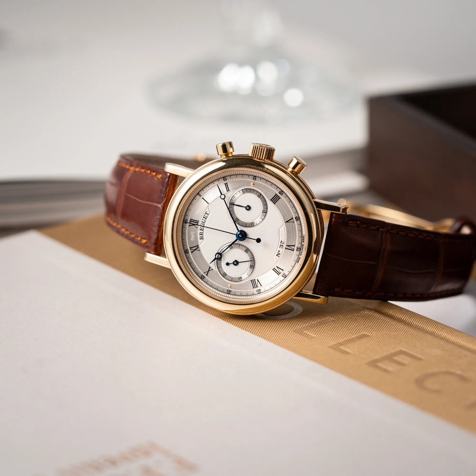 Breguet Classique Chronograph der Referenz 5237 in Gelbgold mit silbernem guillochiertem Zifferblatt am braunem Lederband liegt auf der seitlichen Gehäuseflanke