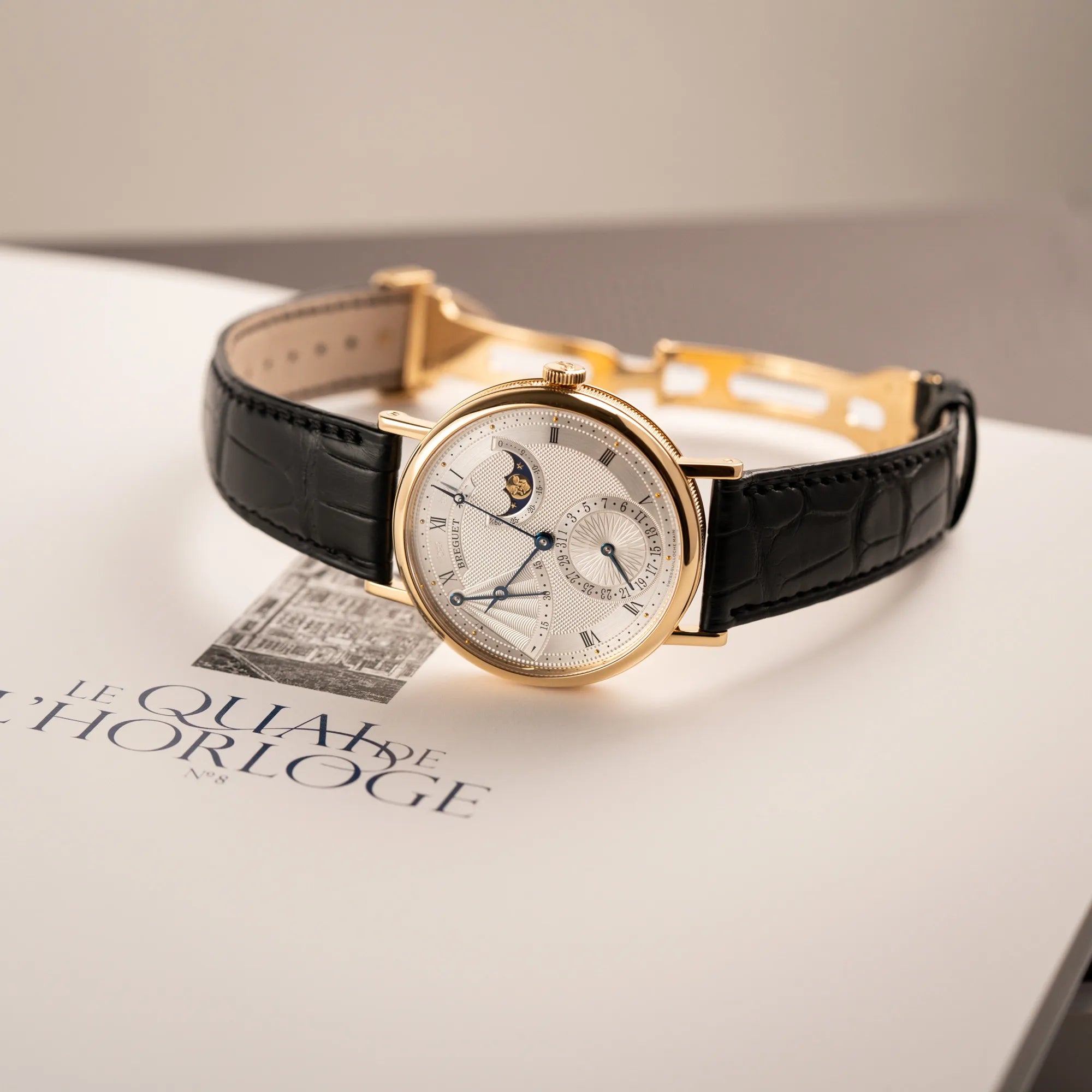 Breguet Classique Mondphase 7137 in Gelbgold mit silbernen, verschieden guillochiertem Zifferblatt liegt auf einem Breguet Buch