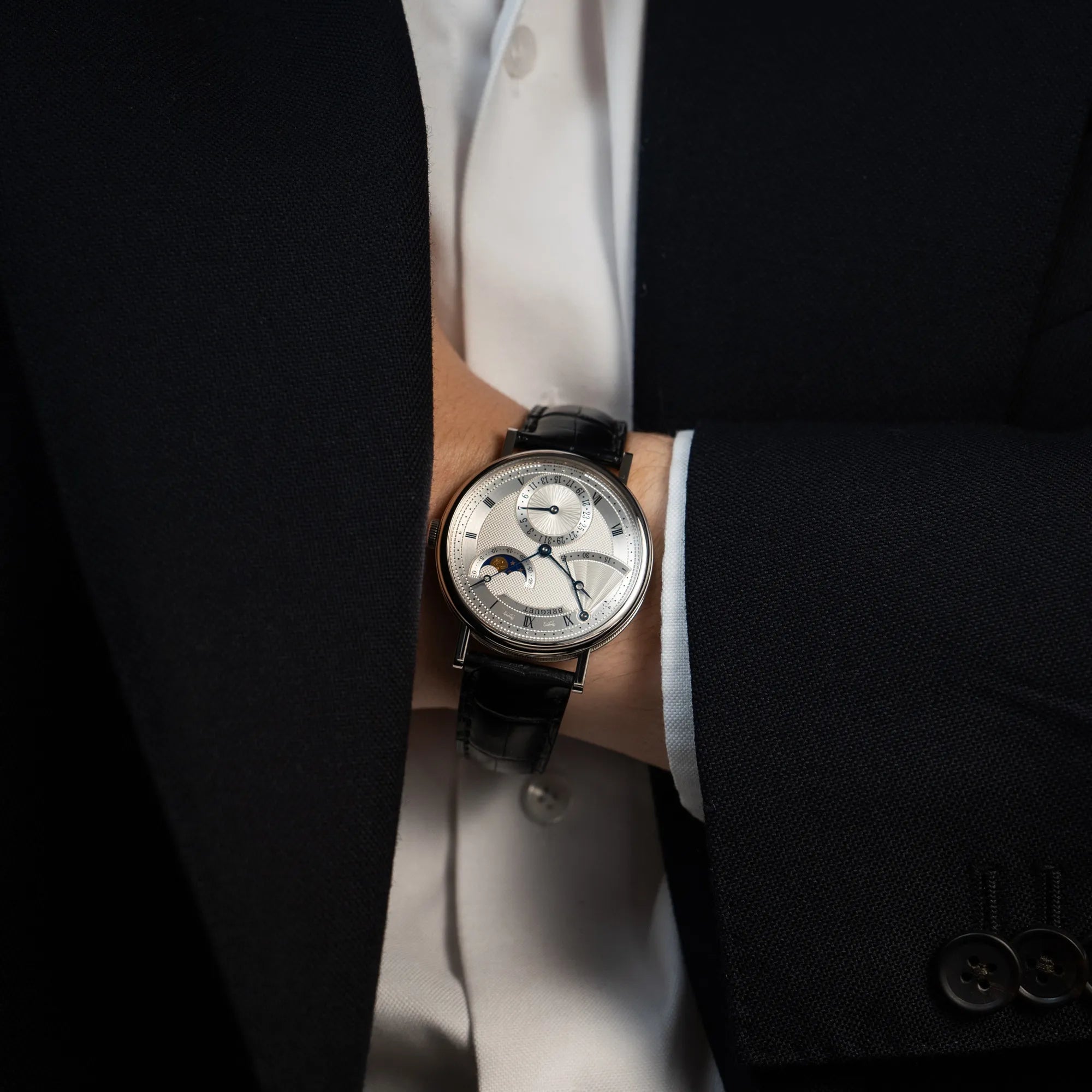 Mann im Anzug hat die Breguet Classique Moonphase der Referenz 7137 in Weissgold mit 3-fach guillochiertem Zifferblatt und Mondphase-Anzeige an seinem Handgelenk