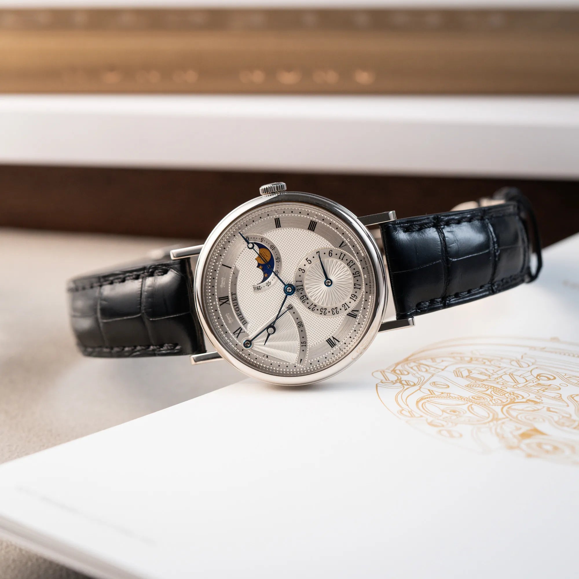 Produktfotografie der seitlich auf dem Gehäuse liegenden Breguet Classique Moonphase der Referenz 7137 in Weissgold mit 3-fach guillochiertem Zifferblatt und Mondphase-Anzeige
