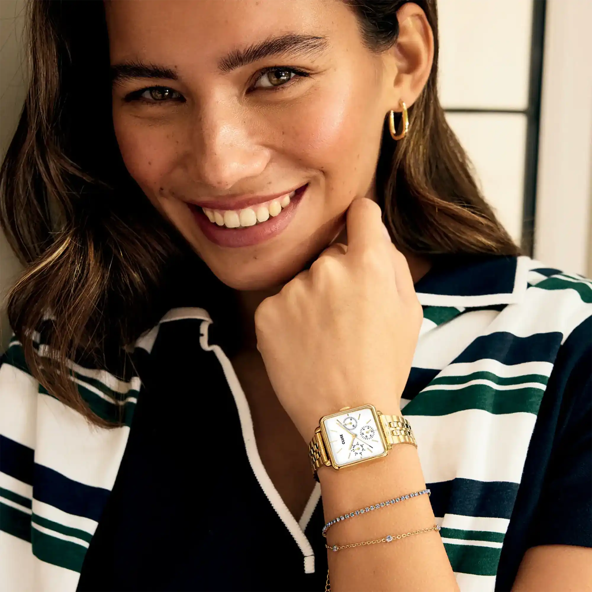 Lifestylebild einer braunhaarigen Frau, die die goldfarbene Cluse Damenuhr La Tetragone Multifunction CW13804 mit weißem Zifferblatt in quadratischem Uhrengehäuse an einem goldfarbenem Edelstahl-Armband an ihrem Handgelenk trägt