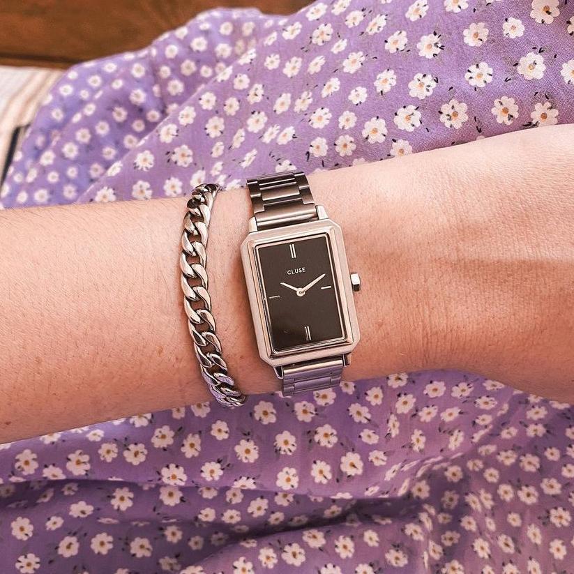 Frau trägt die Cluse "Fluette" Stahl-Uhr mit schwarzem, gestreiften Zifferblatt mit silberfarbenem Gehäuse mit Edelstahl-Armband und kombiniert die Uhr mit silbernen Armbändern - Lila Blumen-Tuch als Hintergrund