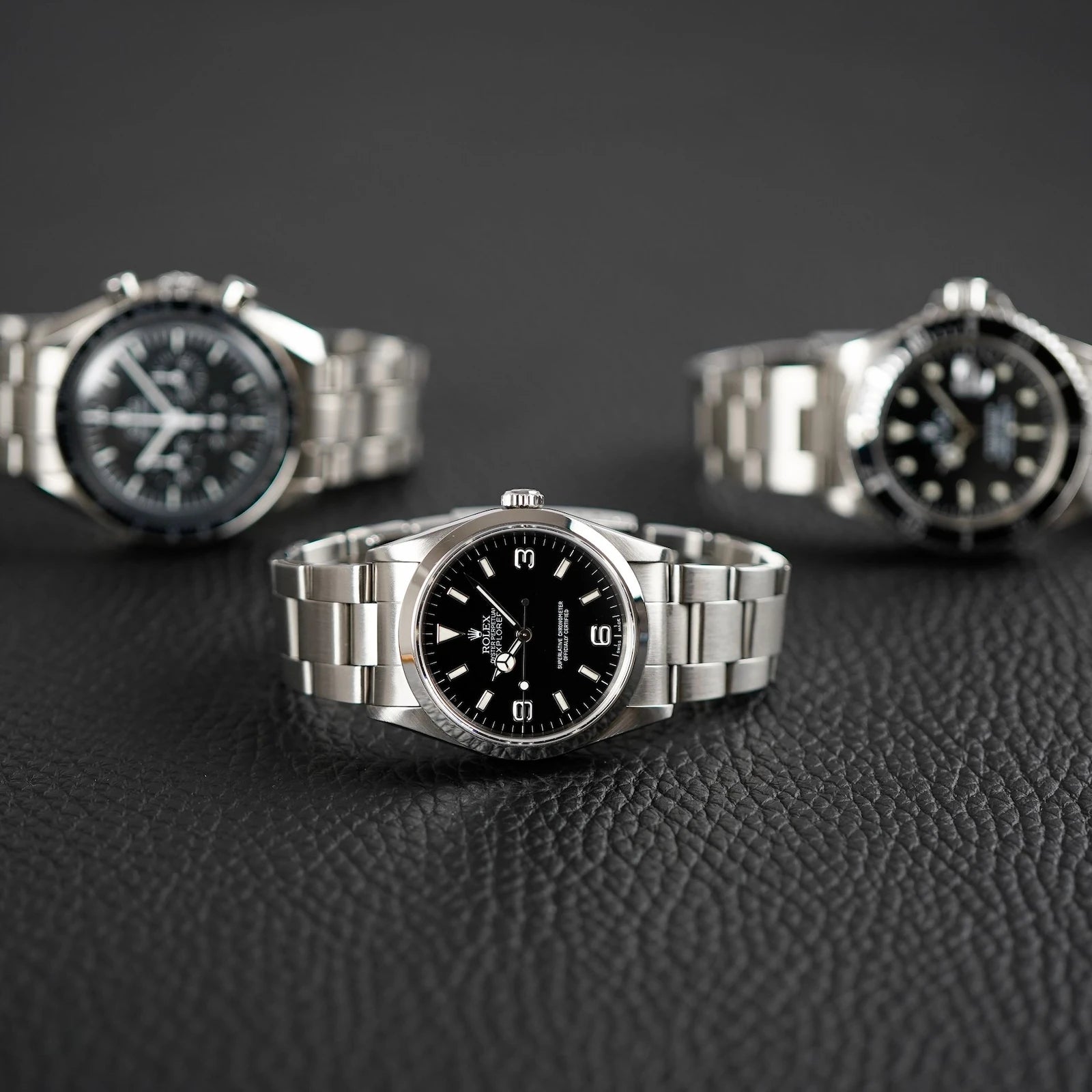 3 Luxusuhren mit schwarzem Zifferblatt liegen auf einer Lederablage - Rolex Explorer, Rolex Submariner & Omega Speedmaster