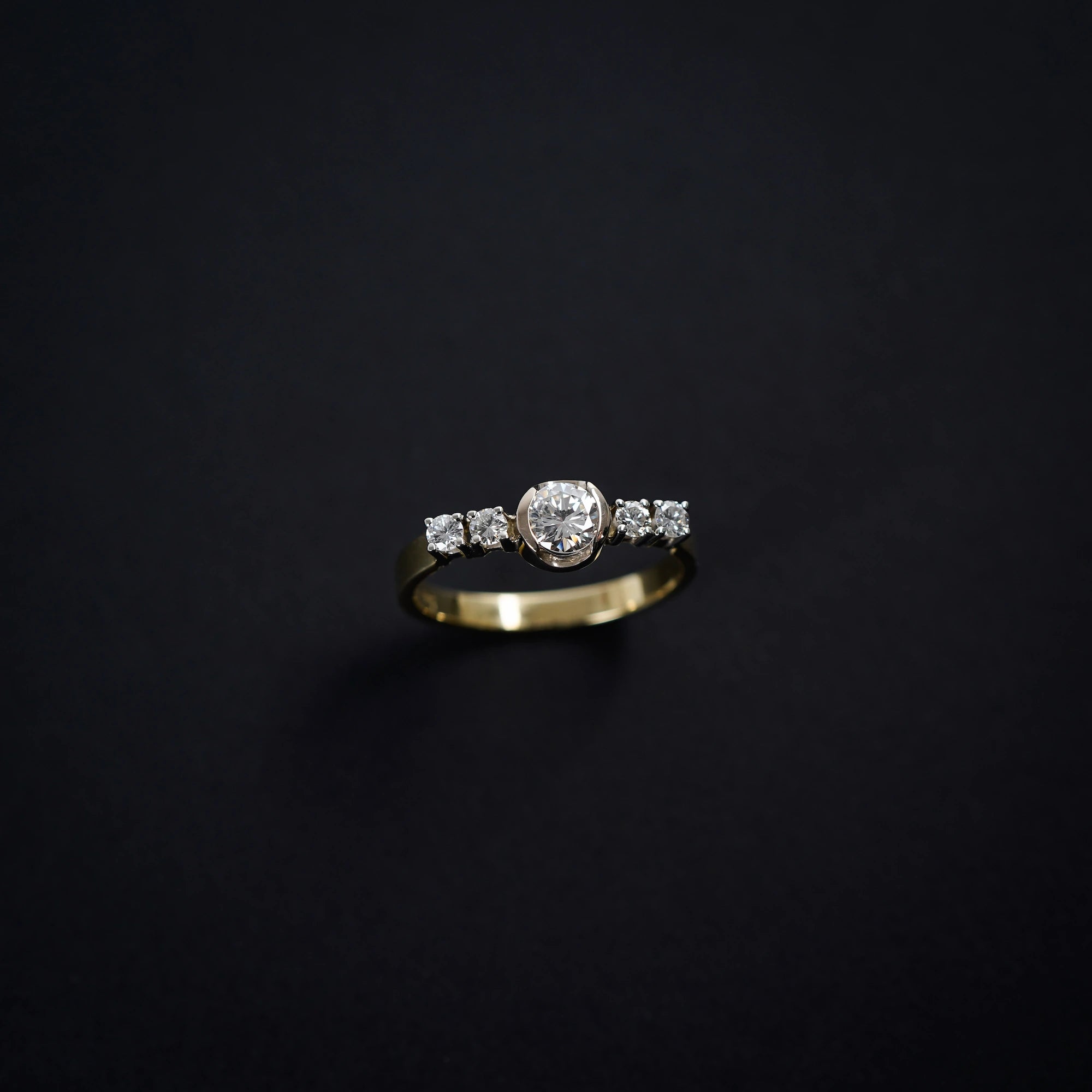 Blick auf auf den gelbgoldenen Ring mit 5 gefassten Diamanten auf der Ringschiene, wovon der zentrale Stein der Größte ist - vor schwarzen Hintergrund