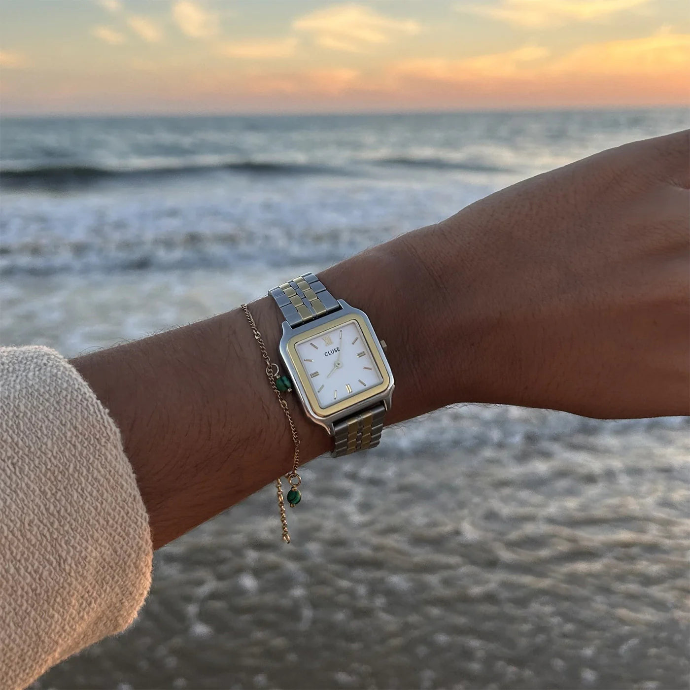 Frau steht am Strand und blickt auf die Frau trägt ein sommerliches Outfit und die Cluse Stahl-Uhr "Gracieuse" in Stahl-Gold-Optik (bicolor) mit weißem Zifferblatt an ihrem Handgelenk