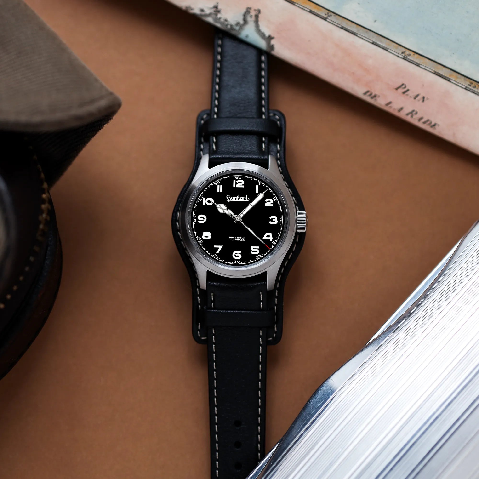 Produktfotografie der Hanhart Preventor HD12 Dreizeiger-Uhr mit schwarzem Zifferblatt am schwarzen Lederband mit Lederunterlage