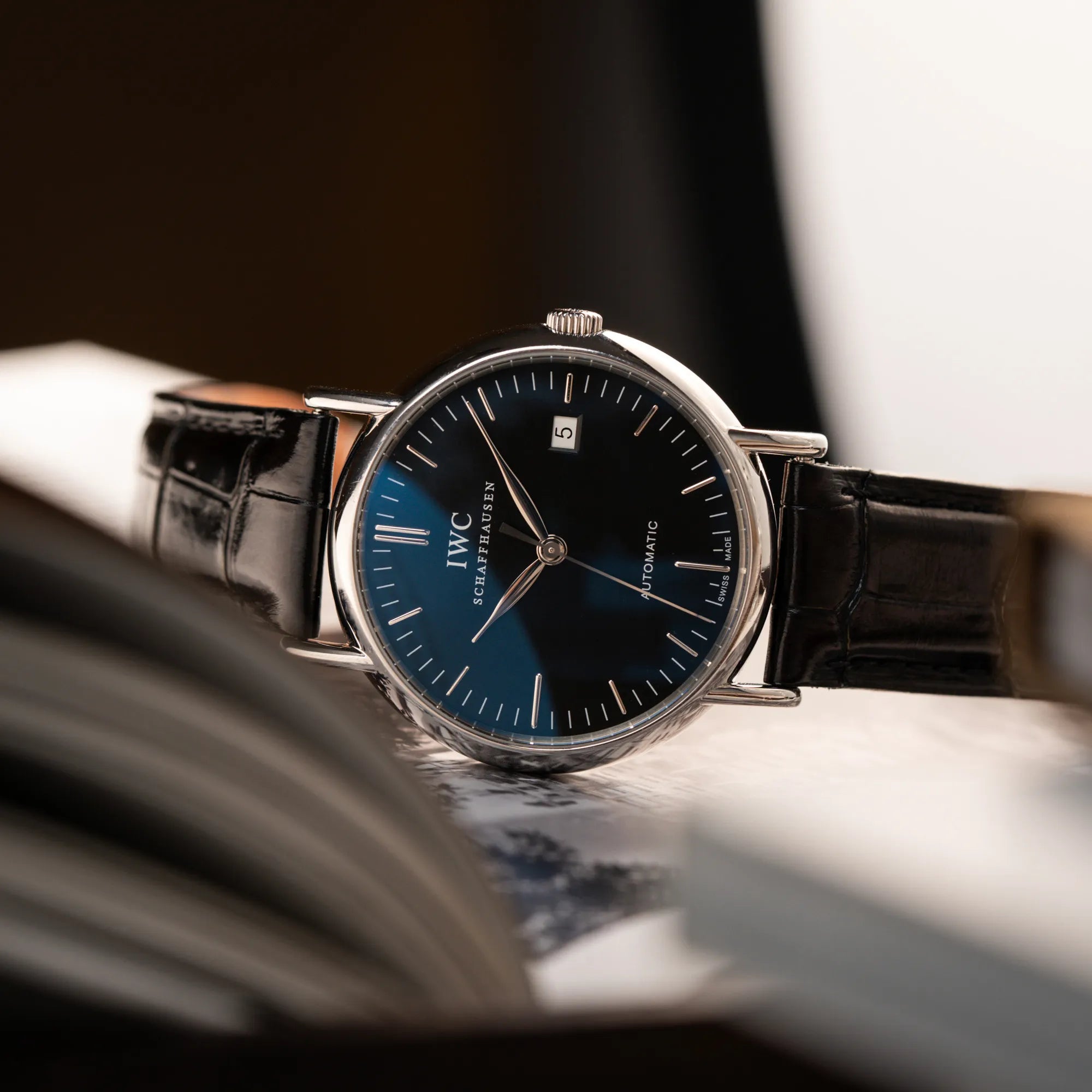 Produktfotografie der seitlich auf der Gehäuseflanke liegenden IWC Portofino Automatik IW356305 Armbanduhr mit minimalistischen, schwarzem Zifferblatt mit Stichindizes am schwarzen Lederband