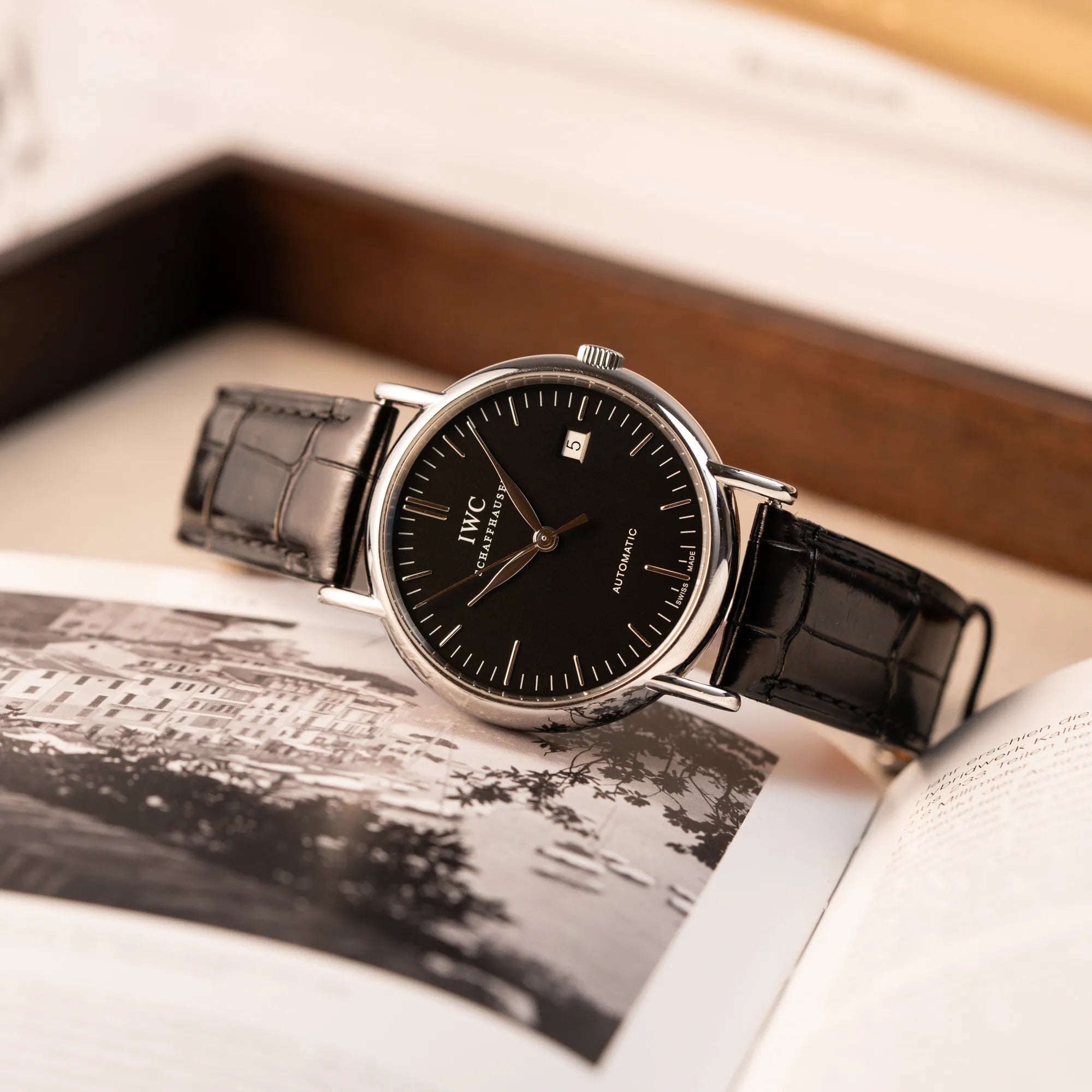 Produktfotografie der IWC Portofino Automatik IW356305 Armbanduhr mit minimalistischen, schwarzem Zifferblatt mit Stichindizes am schwarzen Lederband, während die Uhr auf einem Buch liegt