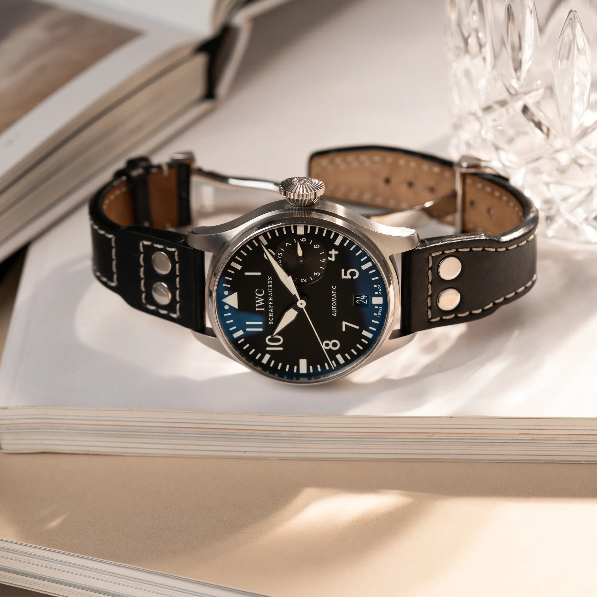 Produktfotografie der IWC Big Pilot 46 Armbanduhr der Referenz IW500401 mit schwarzem Zifferblatt und der Power-Reserve-Anzeige bei 3 Uhr, während die Uhr auf einem Buch liegt