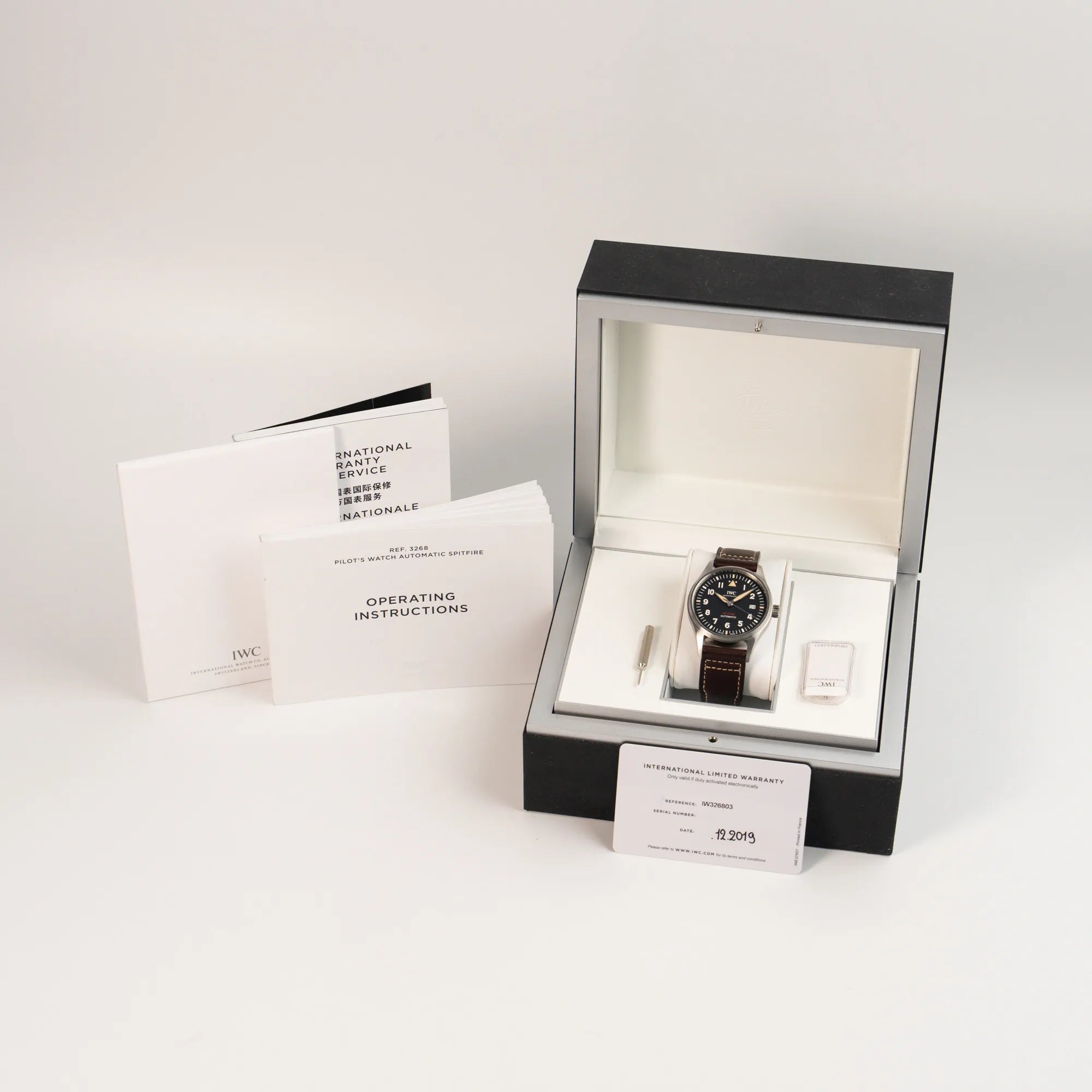 der IWC Pilotenuhr Automatic Spitfire IW326803 mit schwarzen Zifferblatt an einem braunem Lederband zusammen mit dem Lieferumfang, bestehend aus Box, Garantiekarte und Booklets