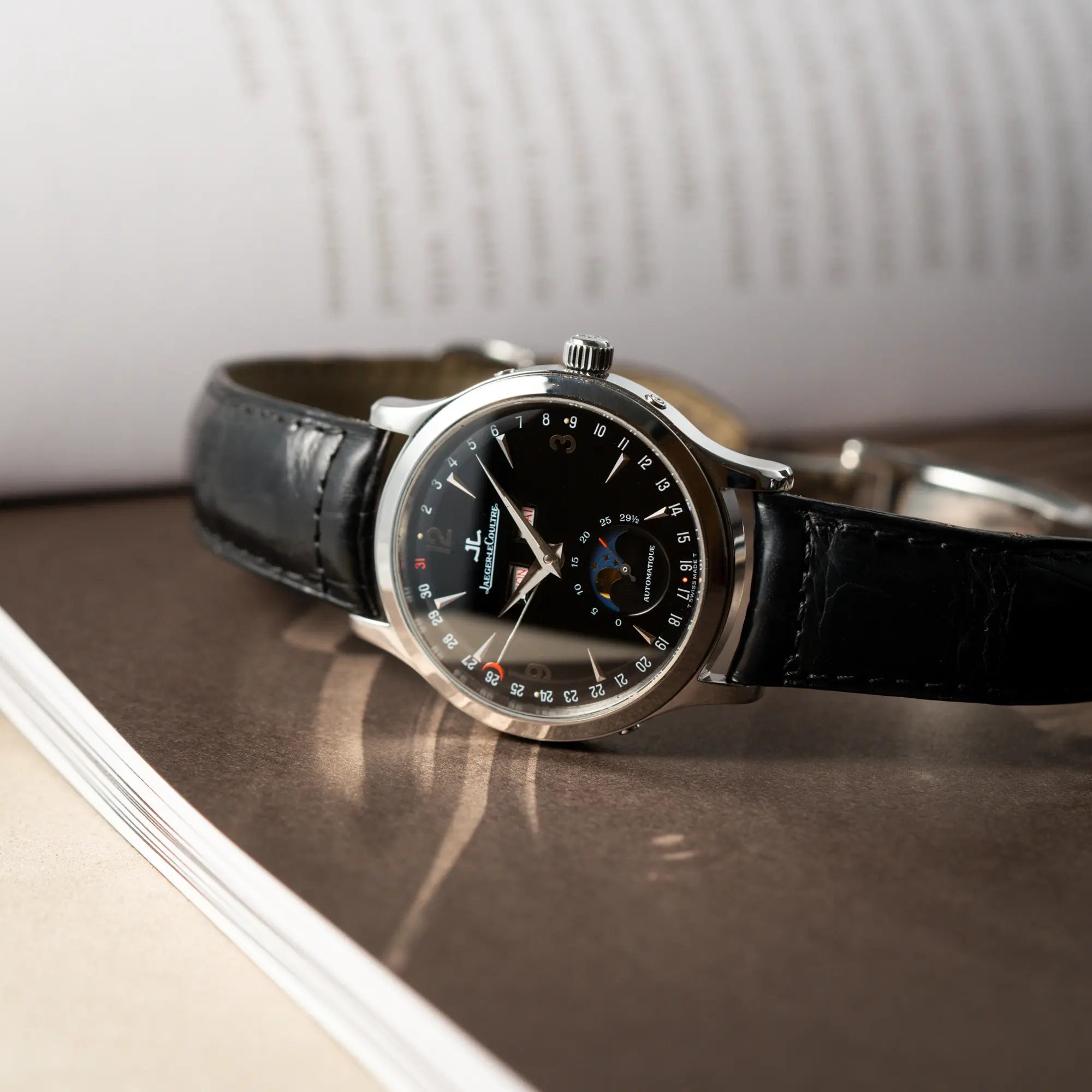 der Jaeger-LeCoultre Master Control Moonphase Calendar mit schwarzem Zifferblatt am schwarzen Lederband liegt seitlich auf dem Uhrenghäuse