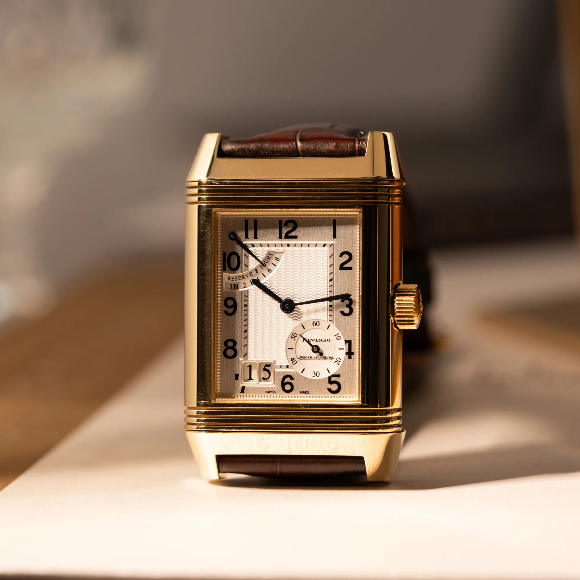 Produktfotografie der Jaeger-LeCoultre Reverso Grande Date der Referenz 240.1.15 mit einem Gelbgold-Gehäuse am braunen Lederband, die senkrecht auf dem Uhrengehäuse steht