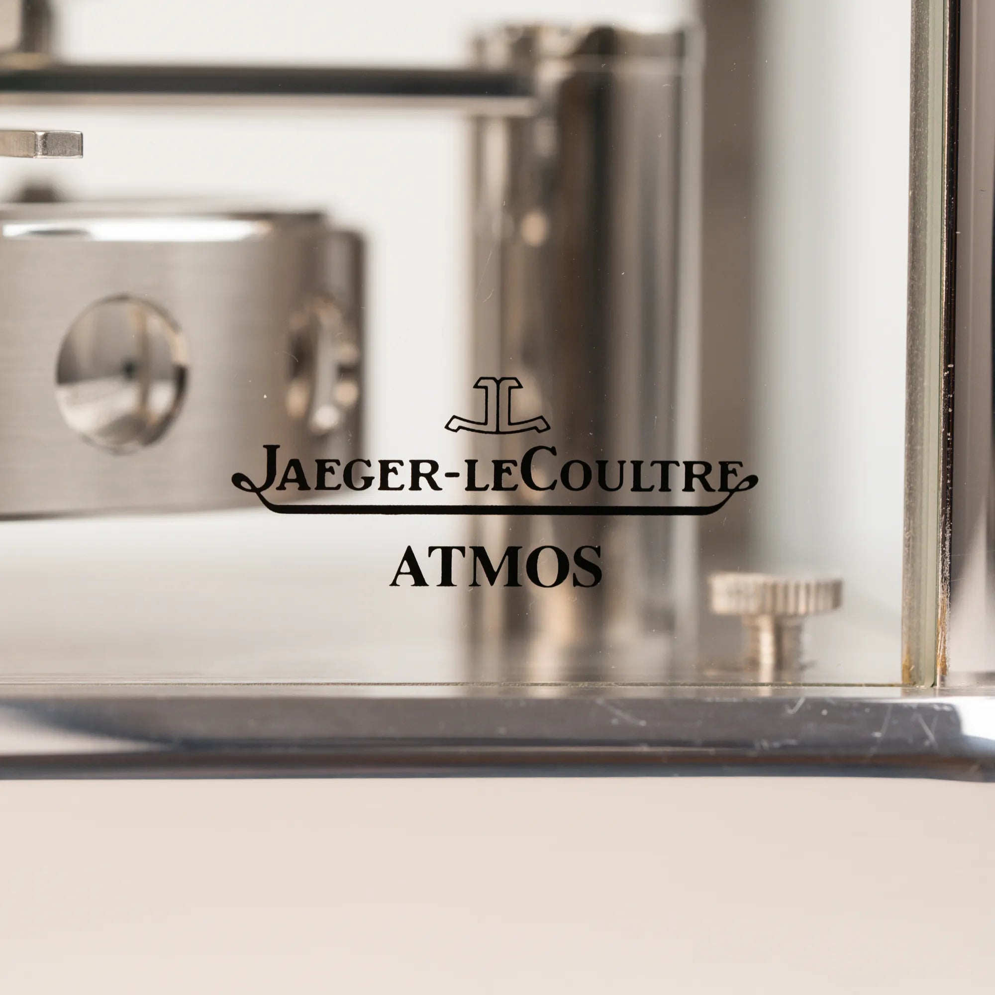 Detailaufnahme des "Jaeger-LeCoultre Atmos" Schriftzugs auf der vorderen Glastür der Jaeger-LeCoultre Atmos Mondphase Tischuhr in rhodiniertem Stahl mit weißem Zifferblatt und blauen römischen Indizes