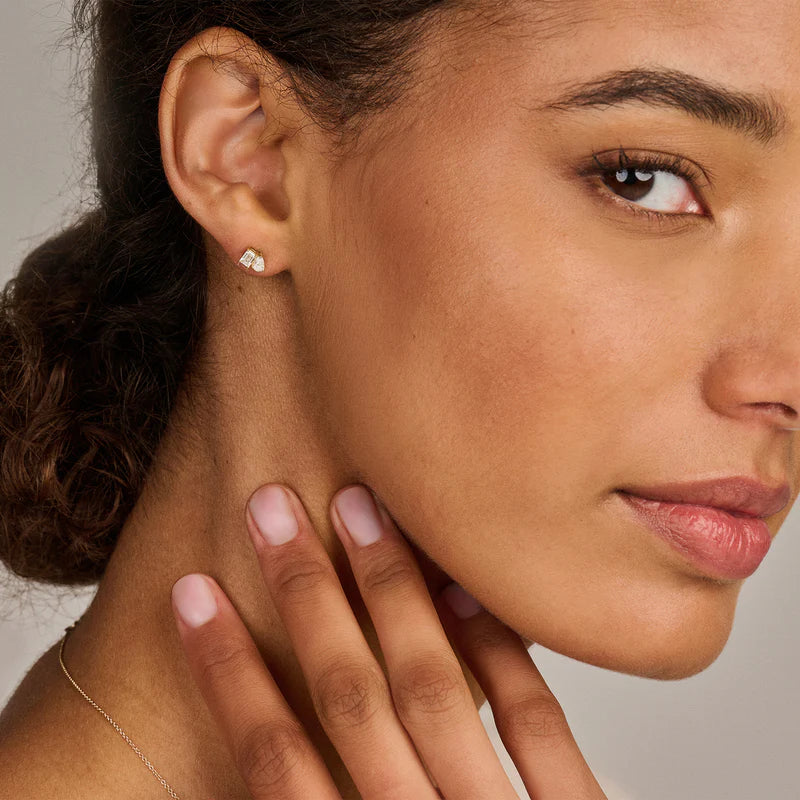 Weibliches Model trägt asymmetrische Ohrstecker aus 14 karätigem Gelbgold mit zwei nebeneinander angeordneten Diamanten, eine Kombination aus einem Diamant im Tropfenschliff und einem rechteckigen Diamant