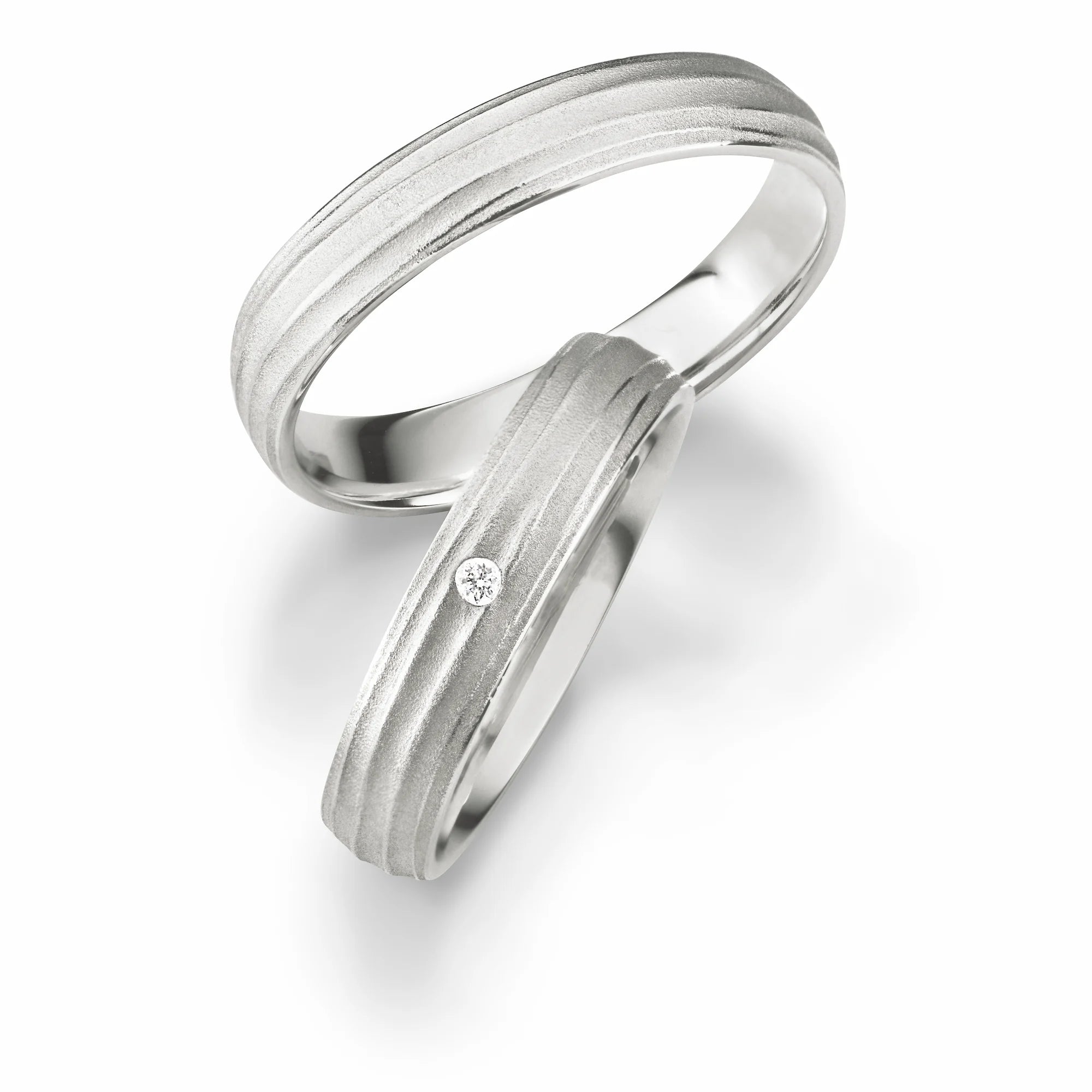 Produktfotografie des weissgoldenen Max Kemper Trauringpaars mit organisch verlaufenden Linien in gebürsteter Oberflächenstruktur und einem zentralen Diamanten als Verzierung des Damenrings