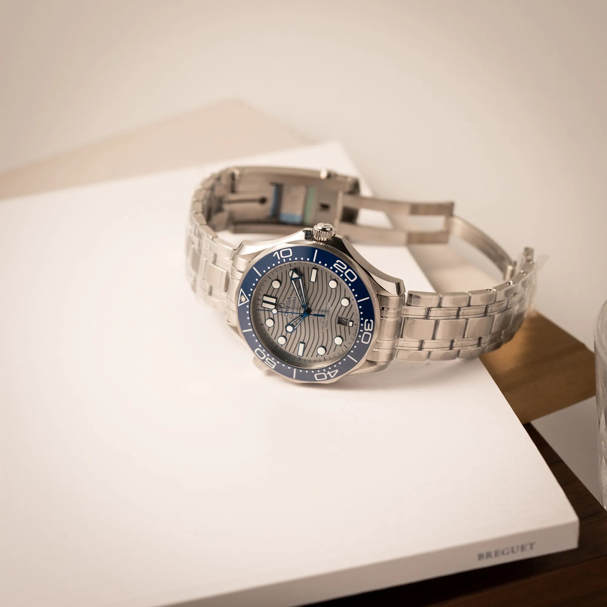 Omega Seamaster Diver 300M der Referenz 210.30.42.20.06.001 mit grauem Zifferblatt im Wellen-Design und blauer Lünette am Edelstahlarmband, welches zum Schutz verklebt wurde, liegt mit geöffneter Faltschliesse auf einem Buch