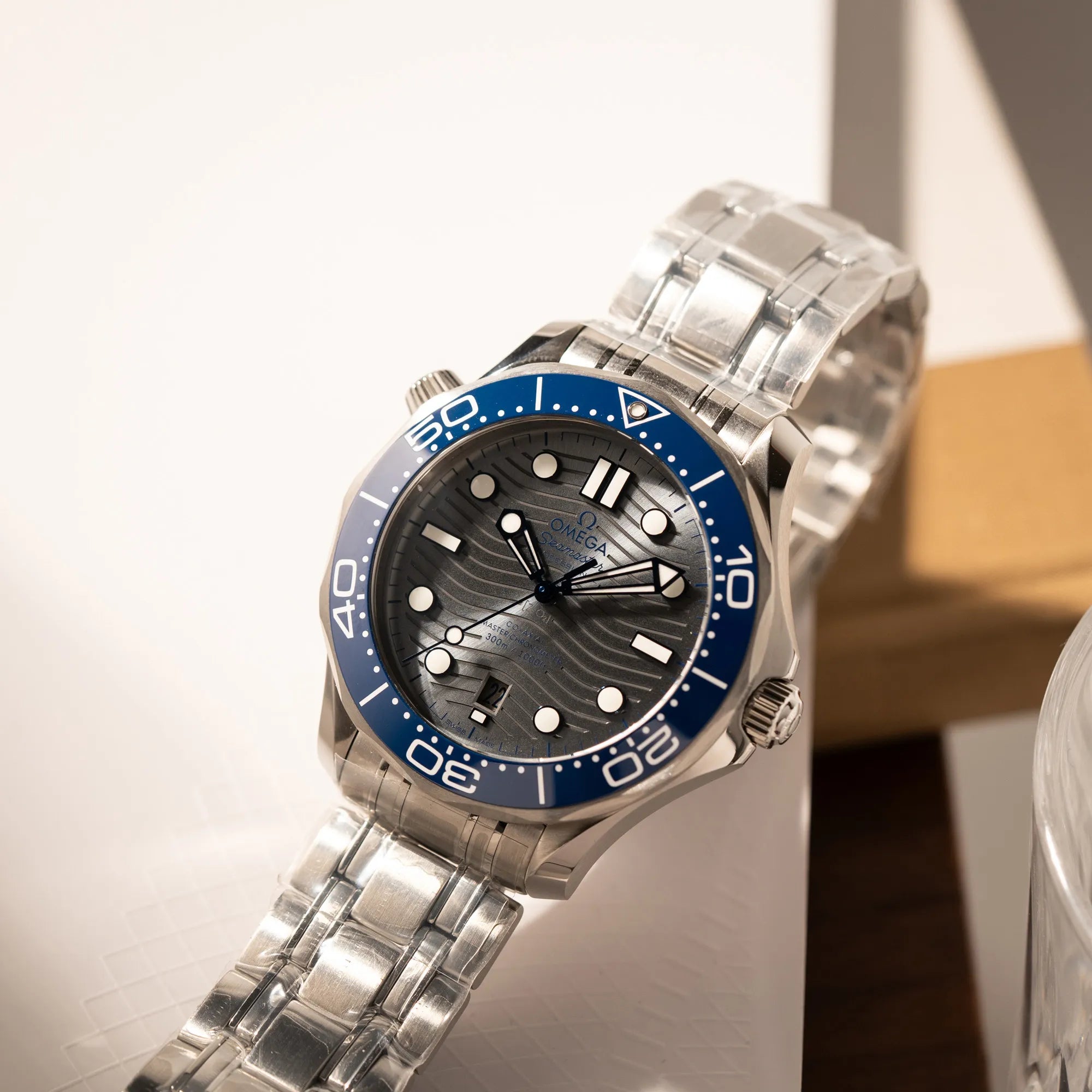 Produktfotografie der Omega Seamaster Diver 300M der Referenz 210.30.42.20.06.001 mit grauem Zifferblatt im Wellen-Design und blauer Lünette am Edelstahlarmband, welches zum Schutz verklebt wurde