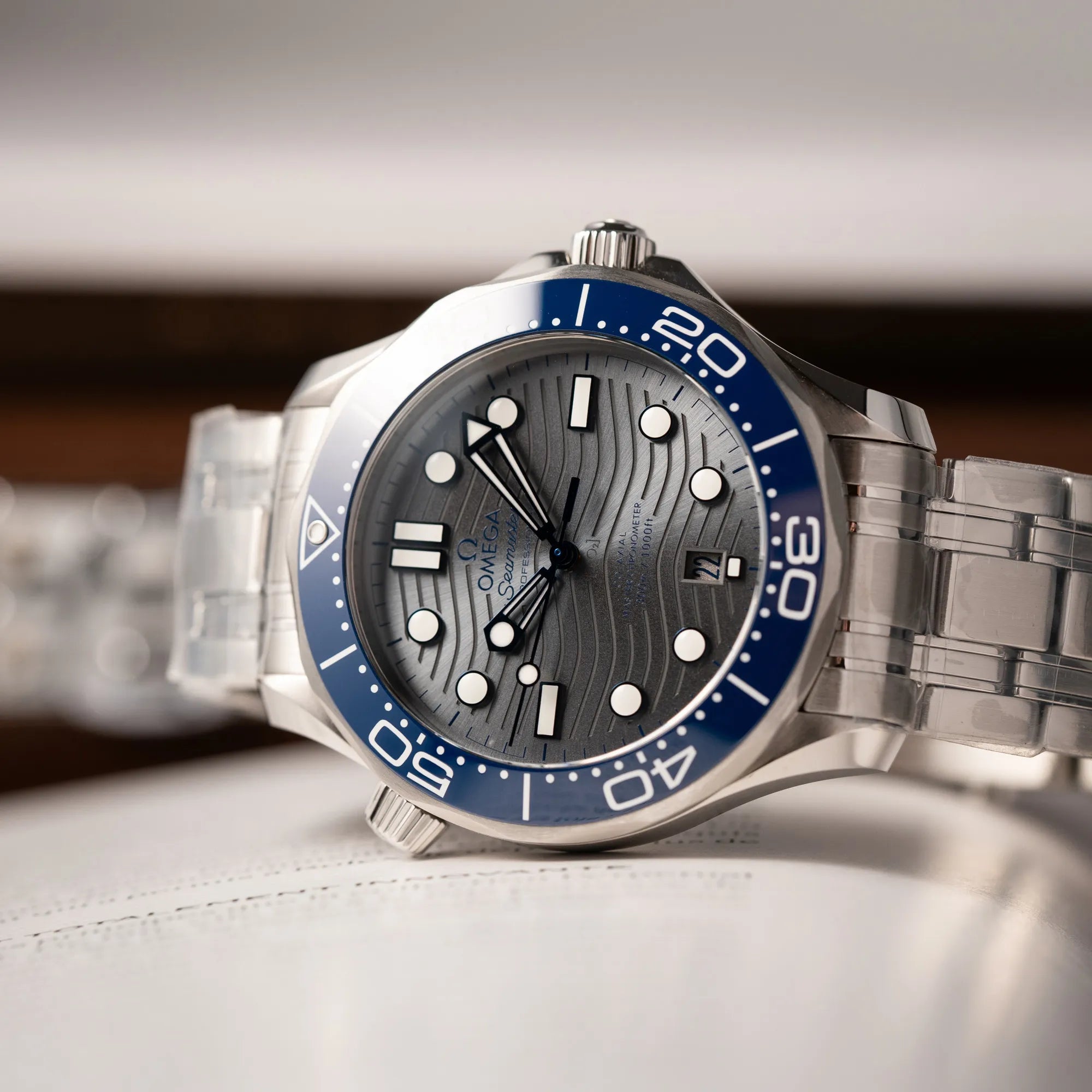 Detailaufnahme der Omega Seamaster Diver 300M der Referenz 210.30.42.20.06.001 mit grauem Zifferblatt im Wellen-Design und blauer Lünette am Edelstahlarmband, welches zum Schutz verklebt wurde