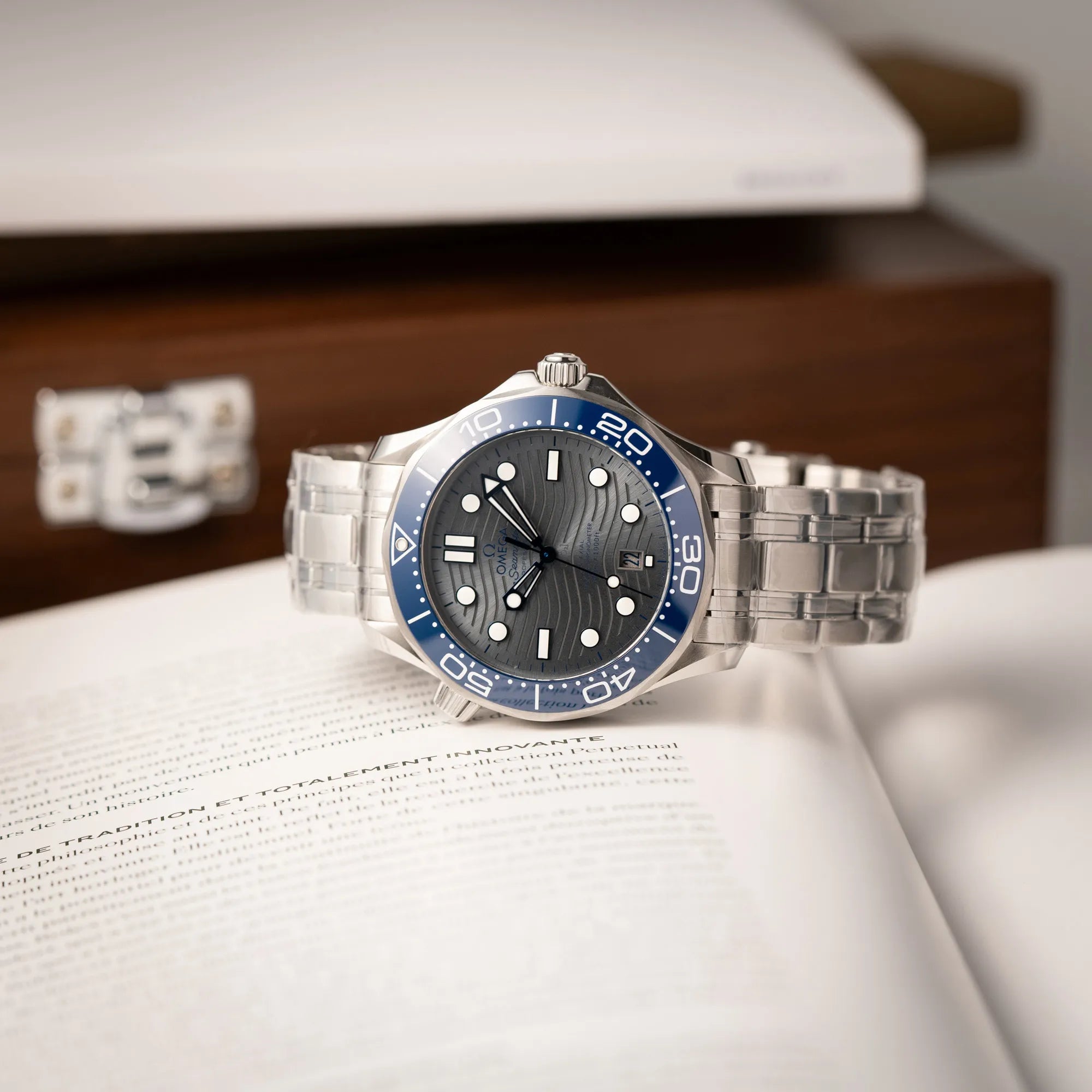 Produktfotografie der Omega Seamaster Diver 300M der Referenz 210.30.42.20.06.001 mit grauem Zifferblatt im Wellen-Design und blauer Lünette am Edelstahlarmband, welches zum Schutz verklebt wurde, liegt auf dem Helium-Ventil