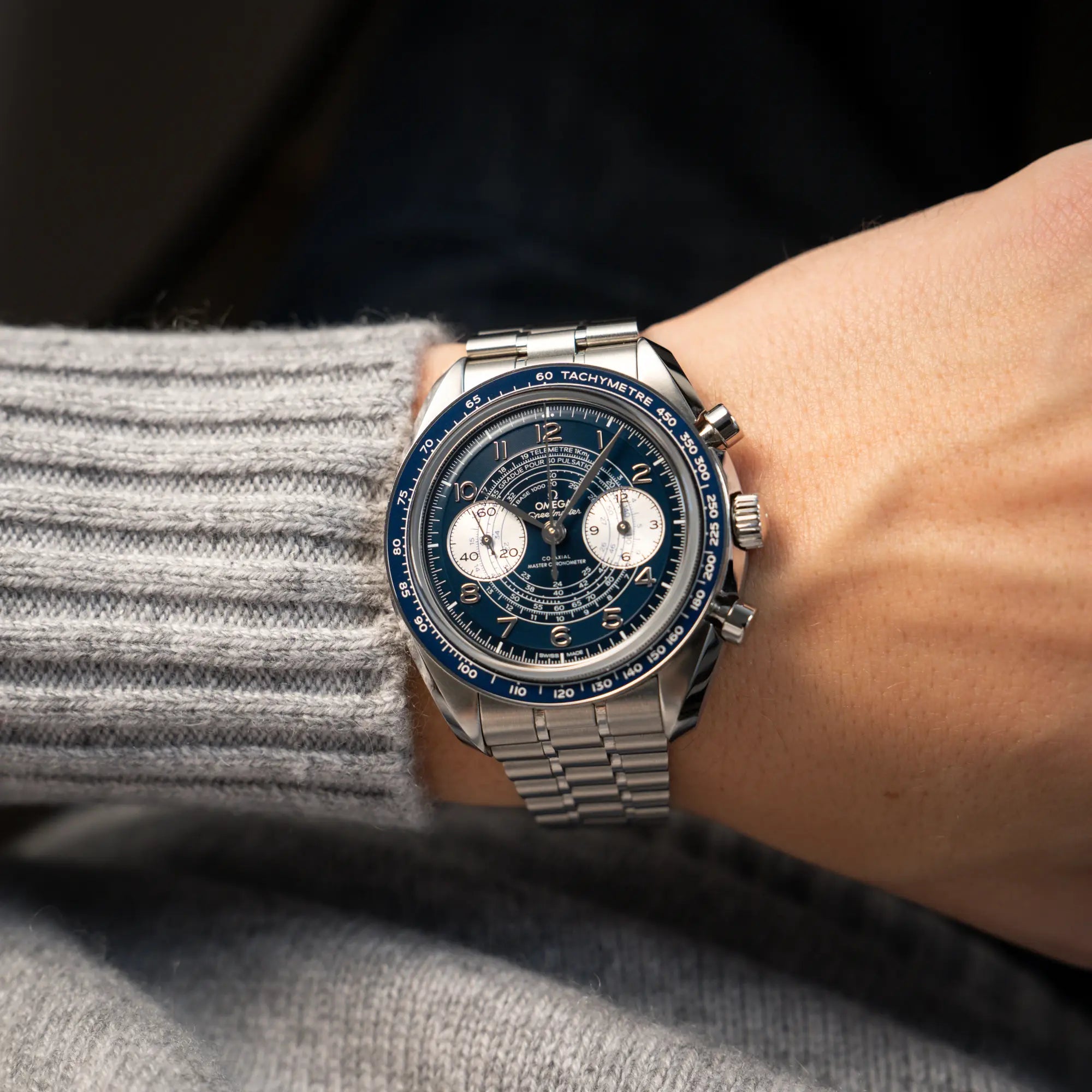 Mann in grauem Poller hat die Omega Speedmaster Chronoscope aus Edelstahl mit blauem Zifferblatt und Pulsometerskala, sowie Telemeterskala an seinem Handgelenk