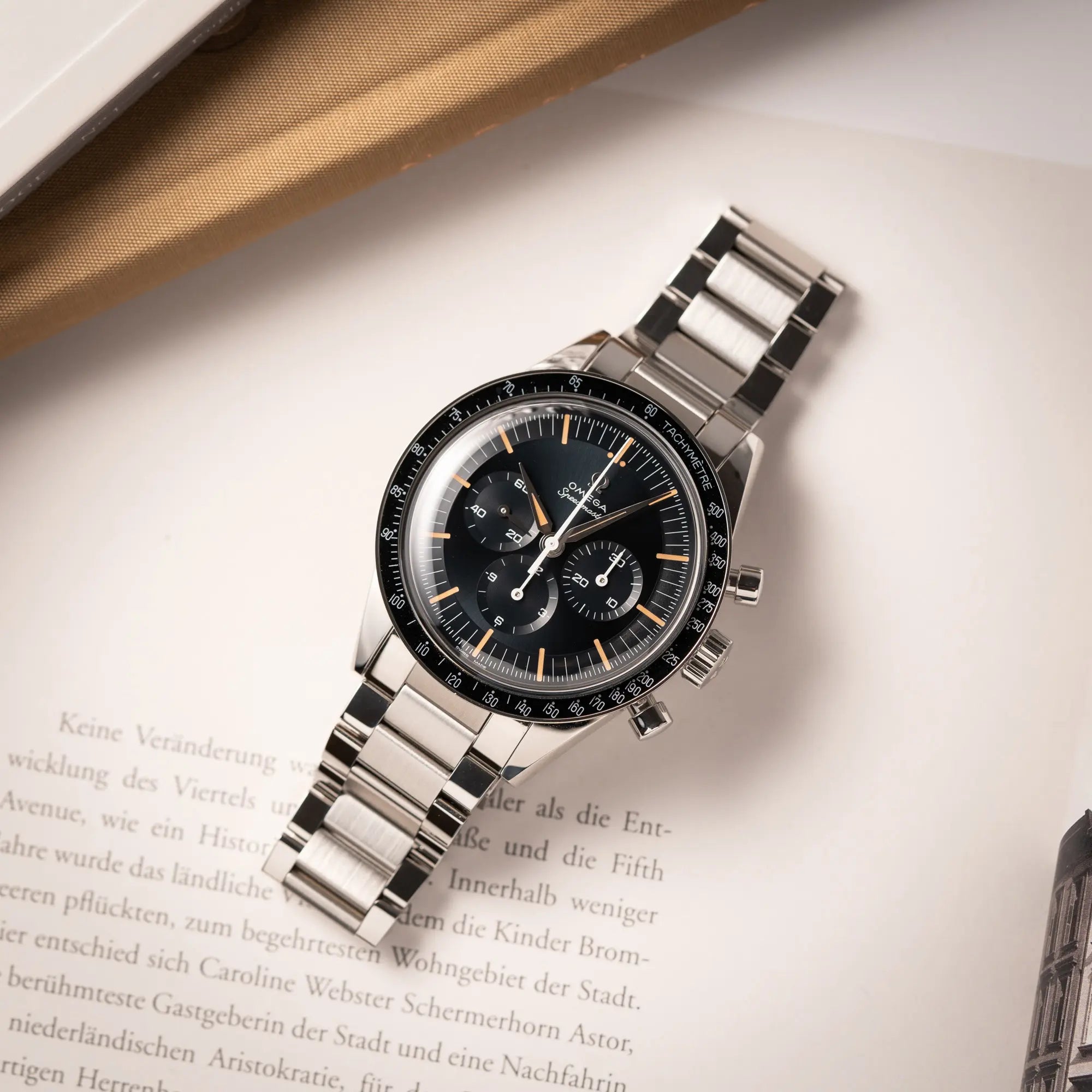 Produktfotografie der flach auf einem Buch liegenden Omega Speedmaster FOIS der Referenz 310.30.40.50.06.001 mit dem dunkelblauen Zifferblatt mit dunkel-beigen Indizes im Vintage-Look und einem vom Standard-Modell abgeänderten Stahl-Armband, was beides an das klassische Speedy-Modell erinnert