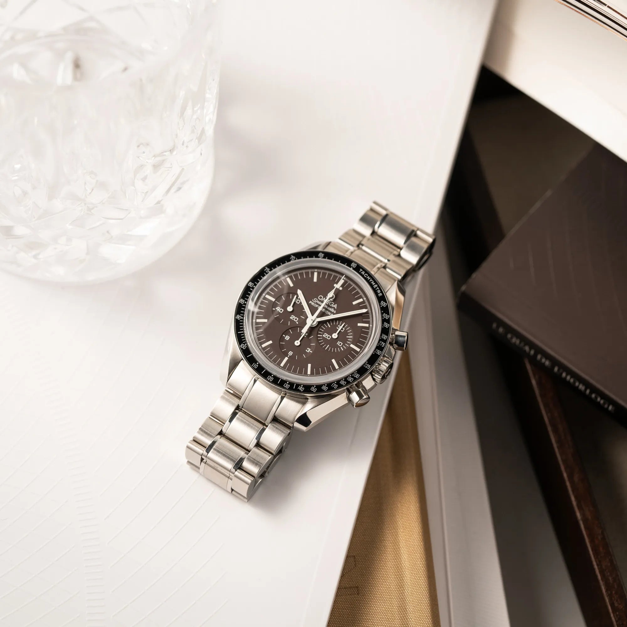 Produktfotografie der flach auf einem Buch liegenden Omega Speedmaster Professional Moonwatch 311.30.42.30.13.001 mit einem braunen Zifferblatt