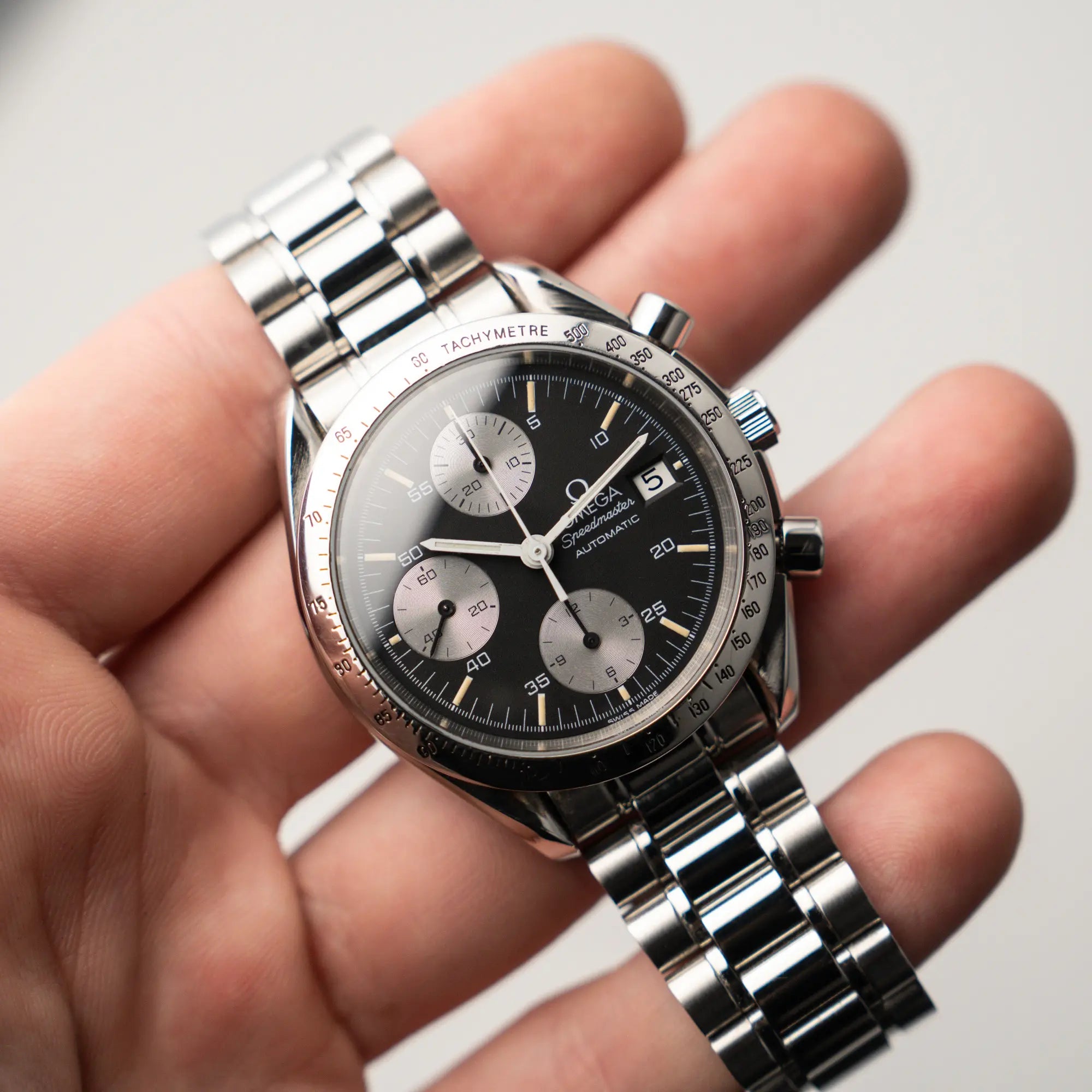Mann hält die Omega Speedmaster Automatic Date mit schwarzem Zifferblatt und silbernen Totalisatoren in seiner Hand und präsentiert die Uhr in die Kamera