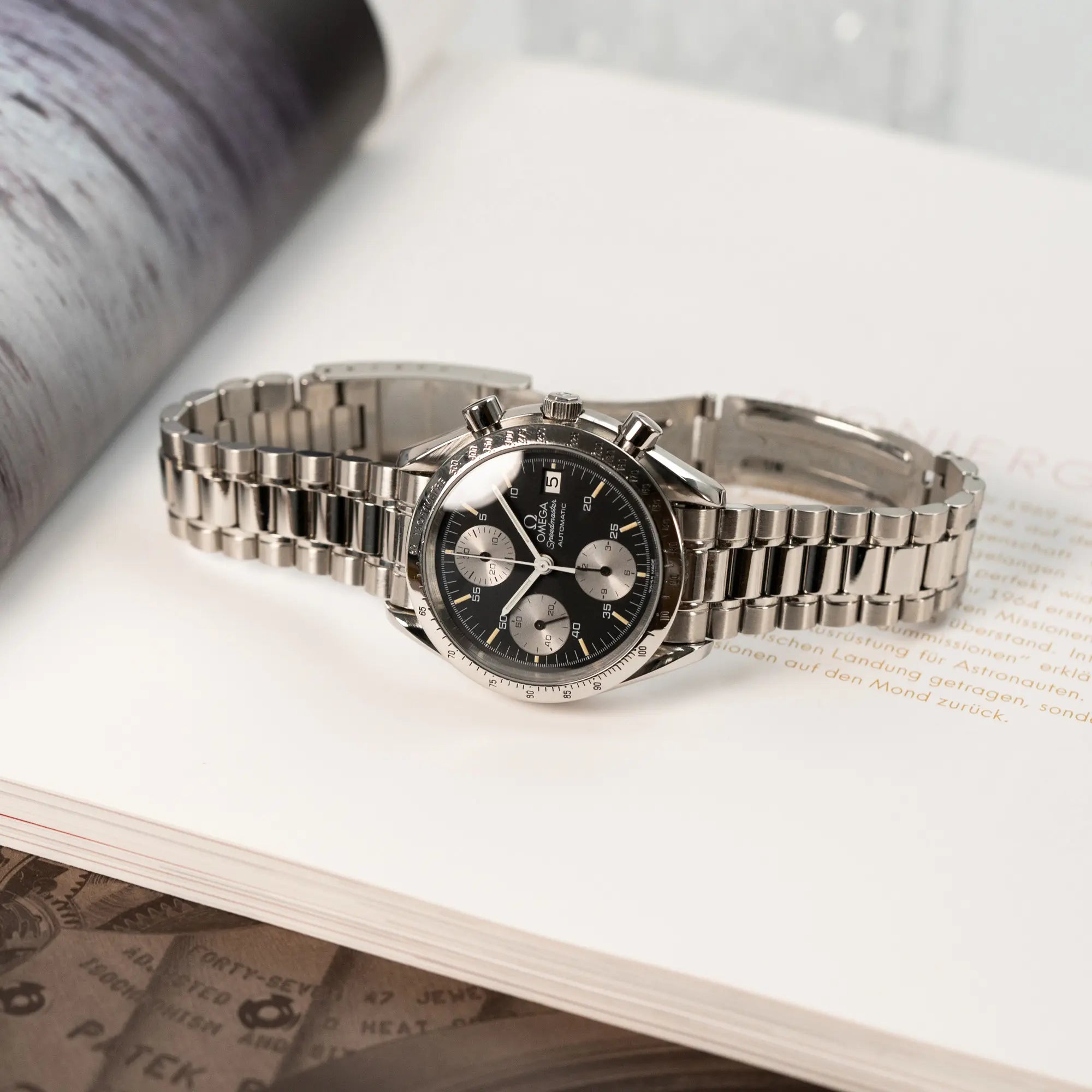Produktfotografie der Omega Speedmaster Automatic Date mit schwarzem Zifferblatt und silbernen Totalisatoren, während die Armbanduhr auf einem Uhrenmagazin liegt