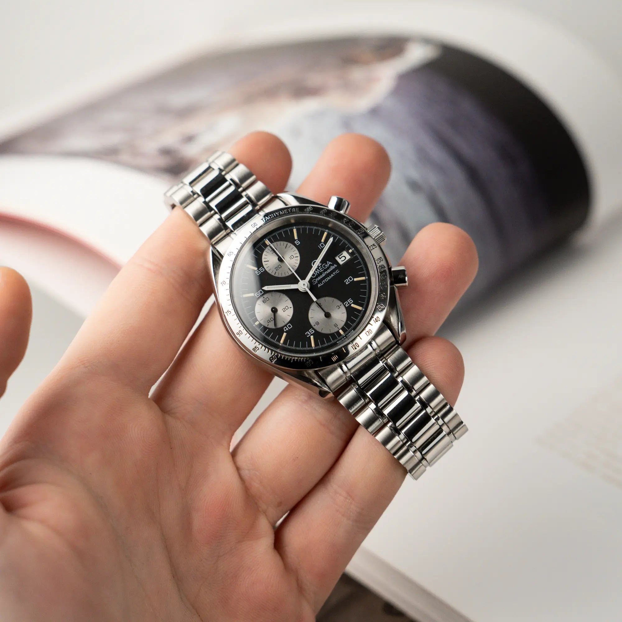 Mann hält die Omega Speedmaster Automatic Date mit schwarzem Zifferblatt und silbernen Totalisatoren in seiner Hand und zeigt die Uhr der Kamera