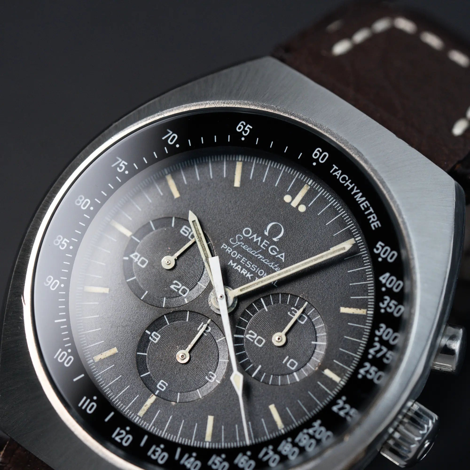 Detailaufnahme des schwarzen Zifferblatts und der Lünette/Gehäuse der Omega Speedmaster Moonwatch Mark 2 an einem braunen Lederarmband