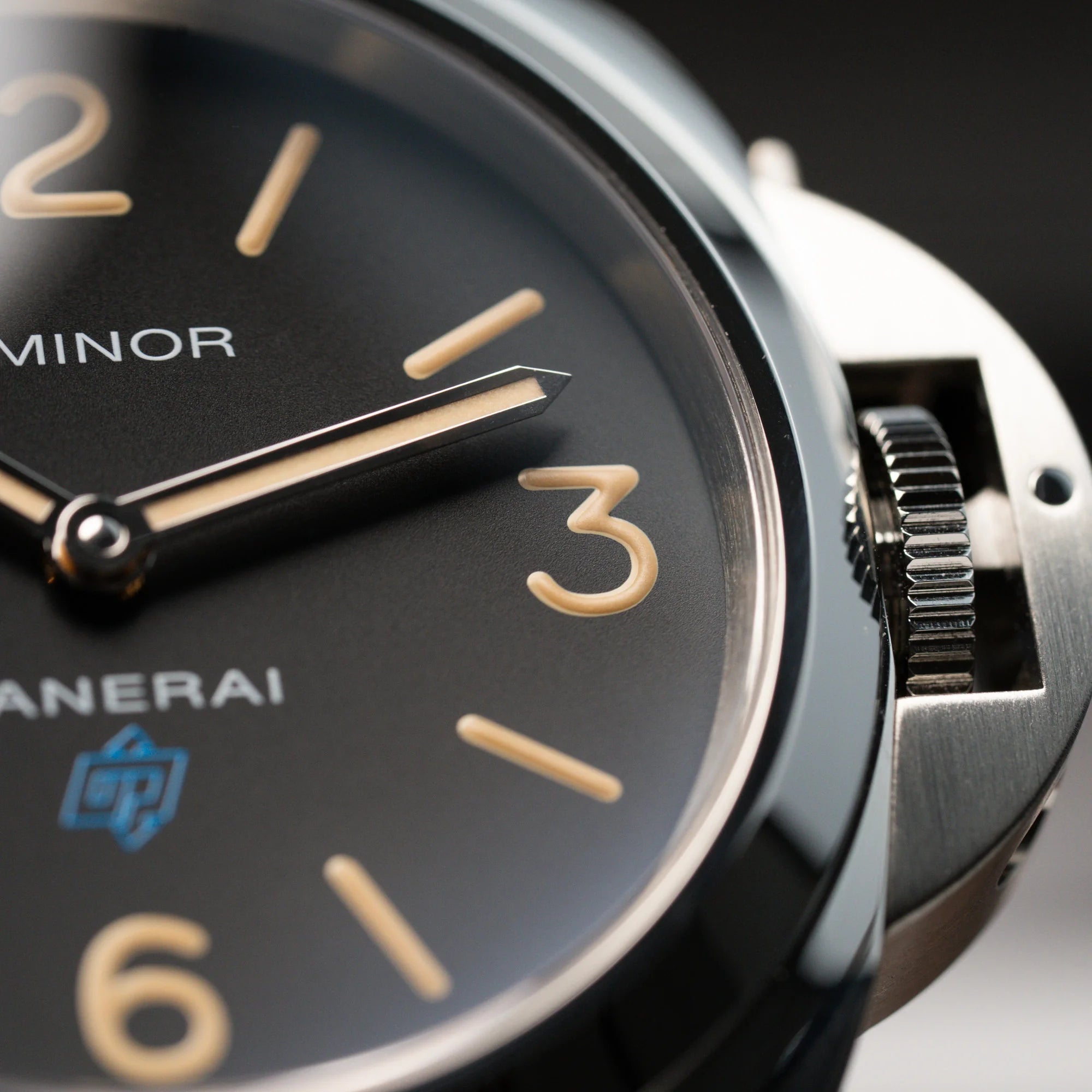 Makroaufnahme mit Fokus auf die Krone und das Zifferblatt der Panerai Luminor Logo Base mit schwarzem Zifferblatt und beigen Indizes, sowie einen schwarzem Lederband