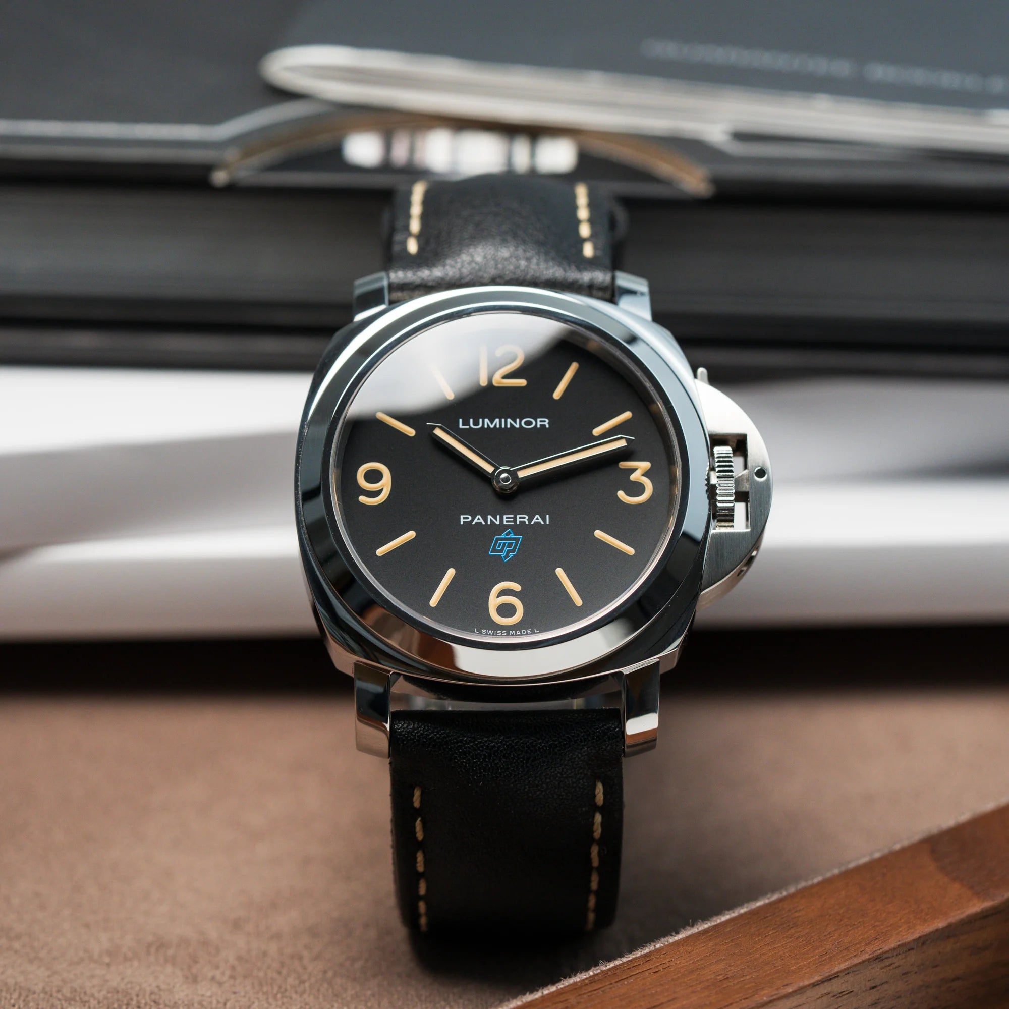 Produktfotografie der Panerai Luminor Logo Base mit schwarzem Zifferblatt und beigen Indizes, sowie einen schwarzem Lederband vor den Booklets