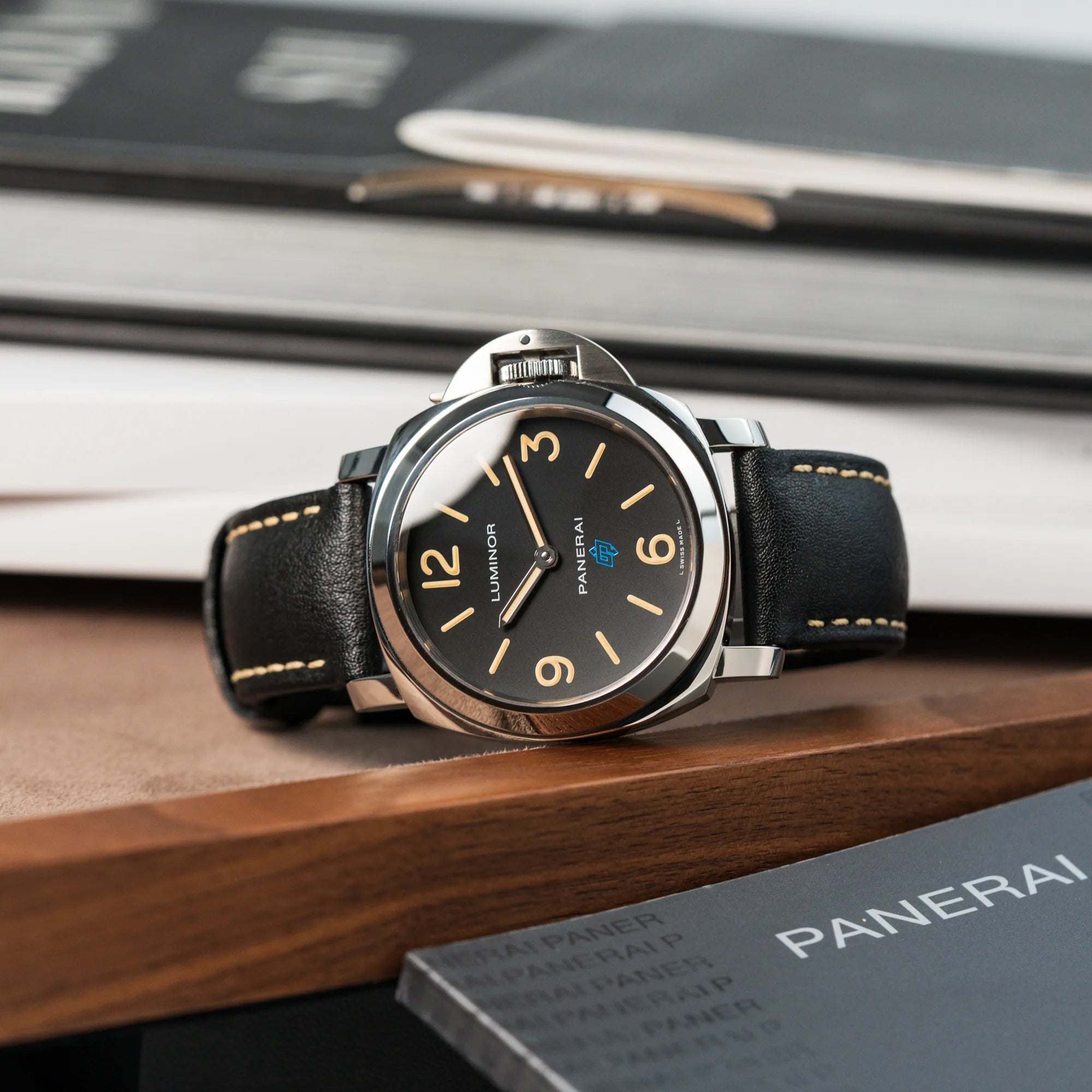 Produktfotografie der liegenden Panerai Luminor Logo Base mit schwarzem Zifferblatt und beigen Indizes, sowie einen schwarzem Lederband vor den Booklets