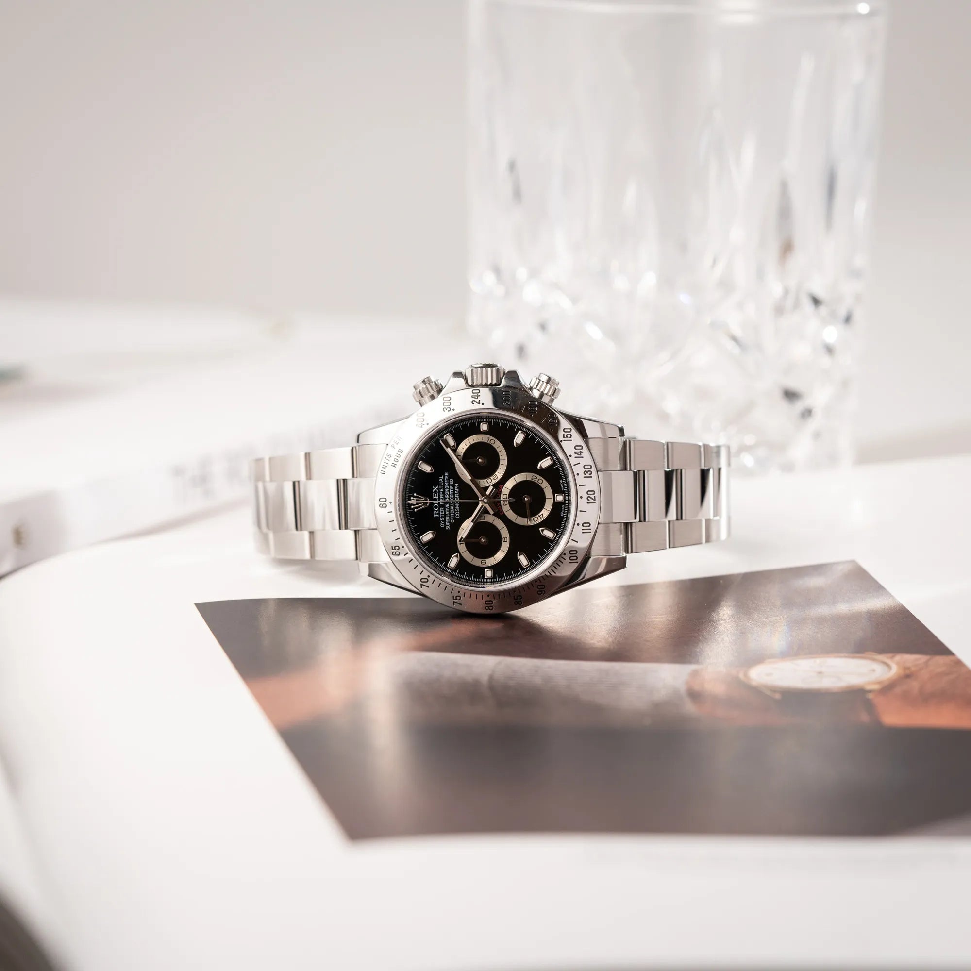 Produktfotografie der auf einem Uhrenbuch liegenden Rolex Cosmograph Daytona 116520 mit schwarzem Zifferblatt und der klassischen Edelstahl-Lünette vor Umstellung auf die Keramik-Lünette