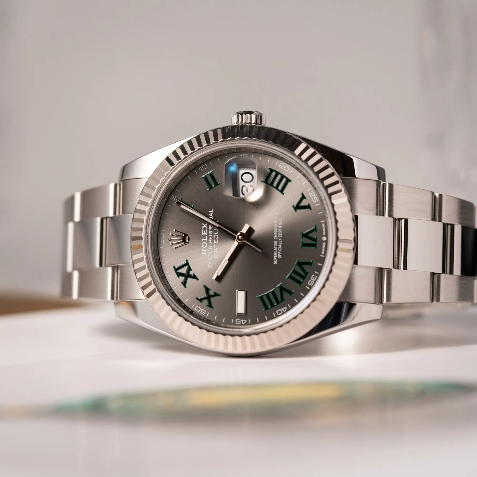 Rolex Datejust 41 der Referenz 126334 mit geriffelter Weißgold-Lünette und Oyster-Stahlband mit dem grauen Wimbledon-Zifferblatt mit schwarz-grünen Indizes liegt seitltich auf dem Uhrengehäuse