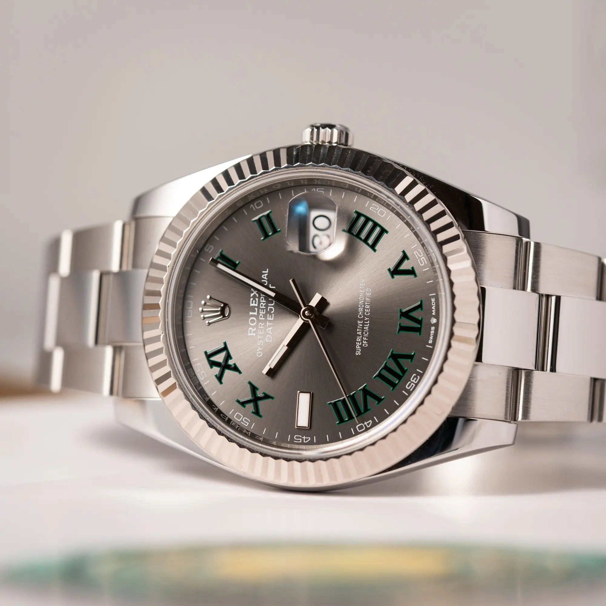 Produktfotografie der auf dem seitlichen Uhrengehäuse liegenden Rolex Datejust 41 der Referenz 126334 mit geriffelter Weißgold-Lünette und Oyster-Stahlband mit dem grauen Wimbledon-Zifferblatt mit schwarz-grünen Indizes