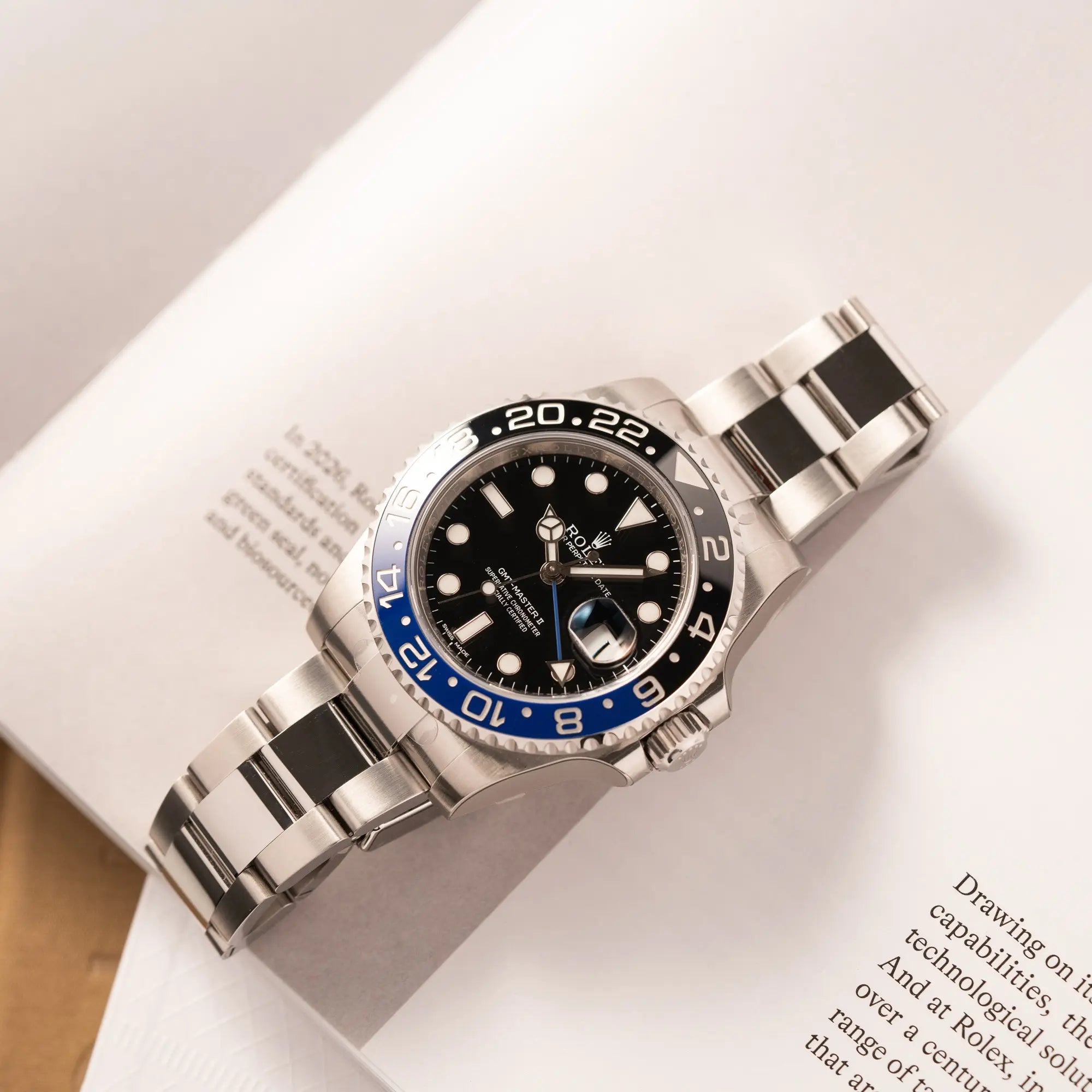 Produktfotografie der flach auf einem Buch liegenden Rolex GMT-Master II der Referenz 116710BLNR mit dem Nickname "Batman" mit der schwarz blauen Keramik-Lünette am Oyster-Stahlband im verklebten Neuzustand