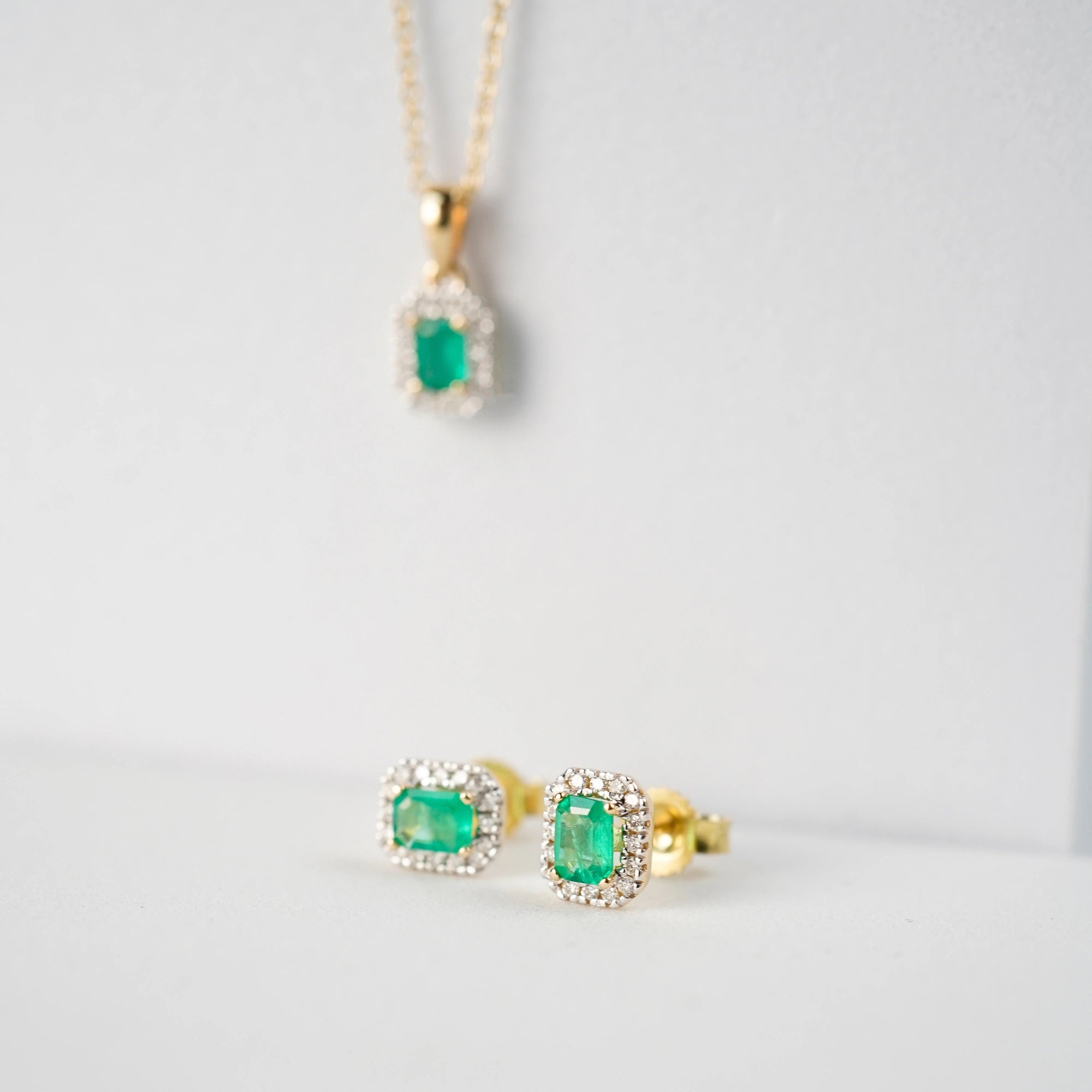 Leicht seitliche Ansicht der aus Gelbgold gefertigten Smaragd-Brillant-Ohrstecker aus der Schmuckatelier Lang Collection, bei denen der zentrale, kräftig grüne Smaragd durch einen Diamantkranz eingerahmt ist, während im Hintergrund unscharf das passende Collier zu erkennen ist.