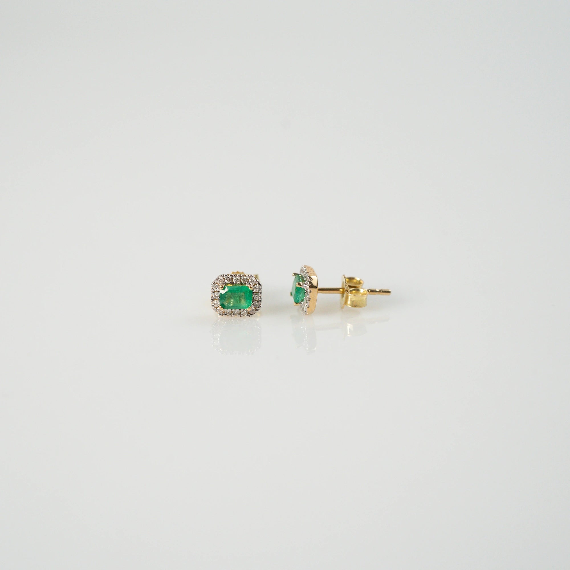Produktfotografie mit zentralem und seitlichem Blick auf die jeweils aus Gelbgold gefertigten Smaragd-Brillant-Ohrstecker aus der Schmuckatelier Lang Collection, bei denen der zentrale, kräftig grüne Smaragd durch einen Diamantkranz eingerahmt ist