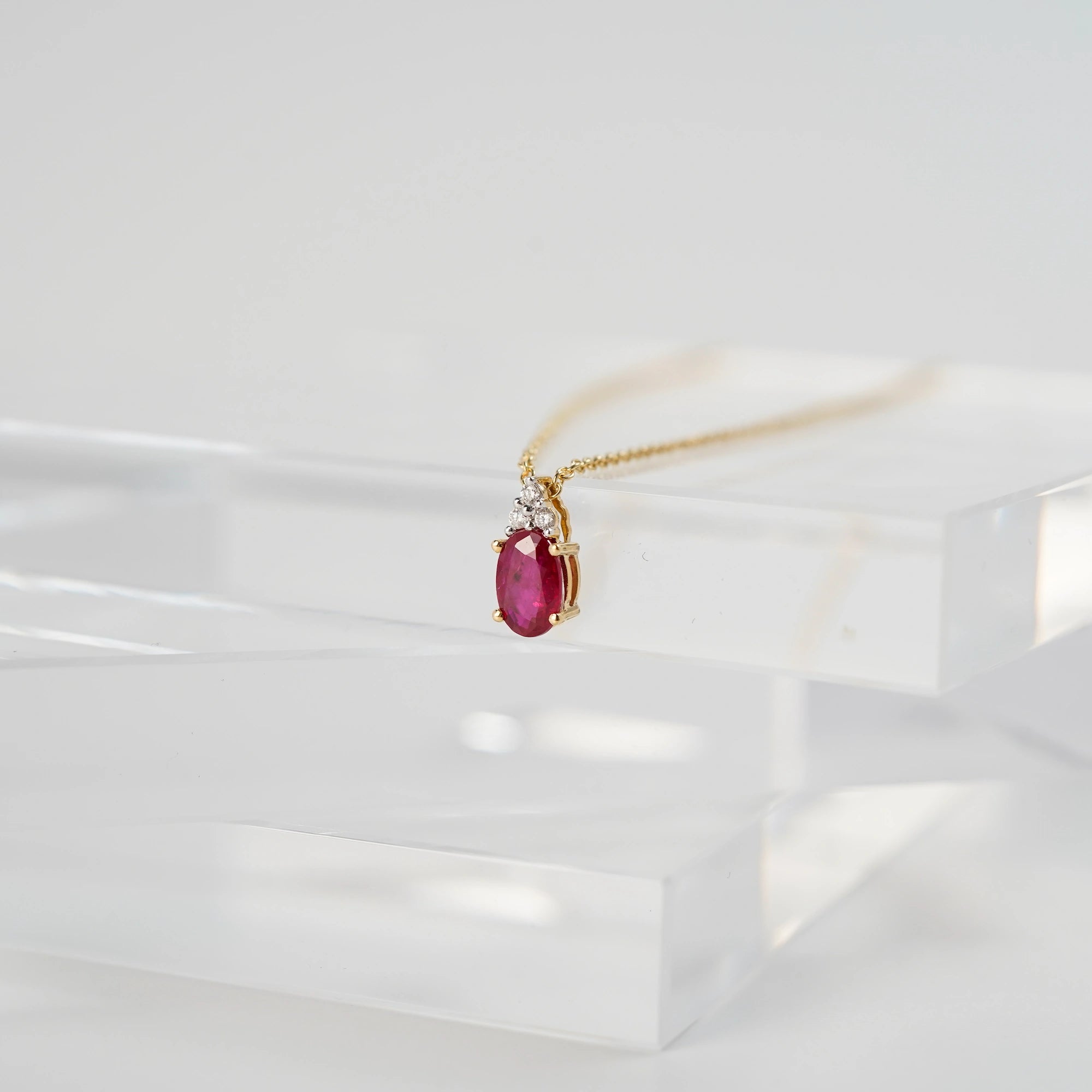 Das Rubin Collier aus der Schmuckatelier Lang Collection an einer gelbgoldenen Kette und einem mit Diamant-verzierten, ovalen Rubin als Anhänger liegt auf transparenten Plastik