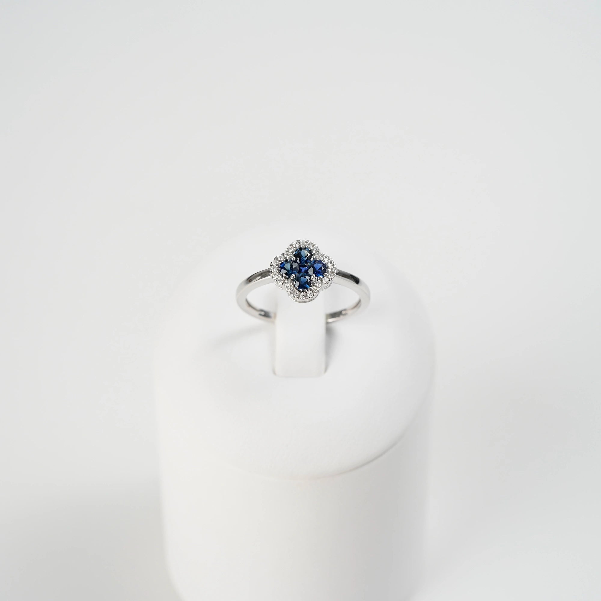 Frontaler Blick auf den Weißgoldring aus der Schmuckatelier Lang Collection mit einer Verzierung in einer Kleeblatt-Form, bei dem die Saphire durch die kleinen Diamanten umrandet sind (auf Ringständer präsentiert)