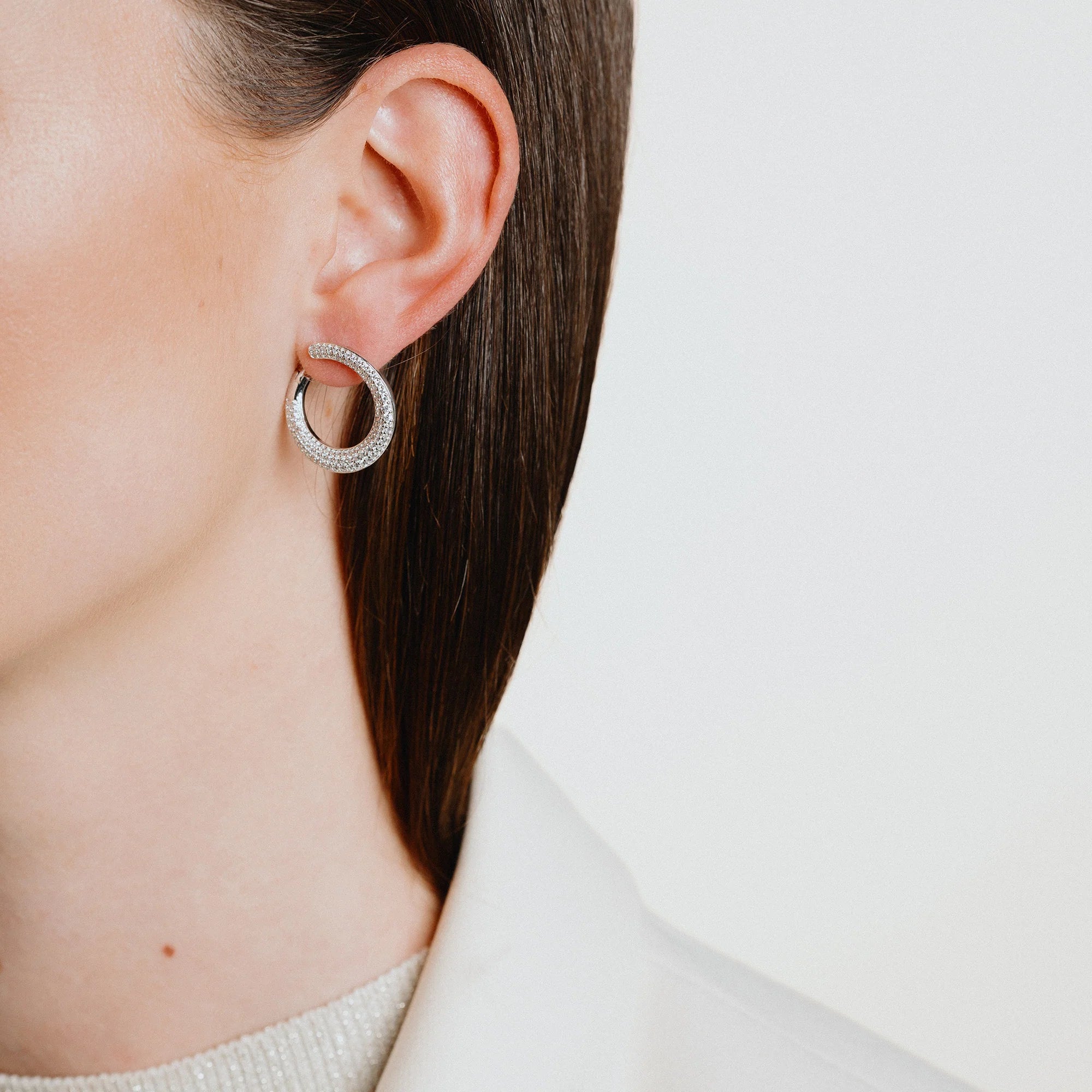 Fotografie der Ohrenpartie einer braunhaarigen Frau, die die Diamonfire-Ohrstecker in einer runden, geschwungenen Form (mit "Öffnung") aus Silber mit einem kompletten Pavé-Besatz, trägt