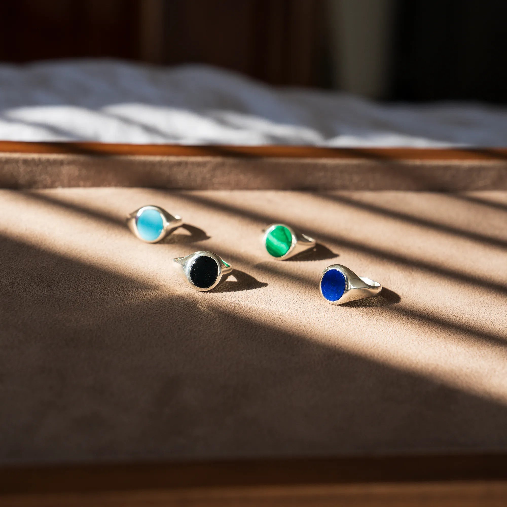 Produktfotografie auf einem braunem Holztablett mit den vier aus Silber handgefertigten Siegelringe aus der Schmuckatelier Lang Collection mit Malachit, Onyx, Türkis oder Lapis Lazuli, während das Licht auf das Tablet einfällt