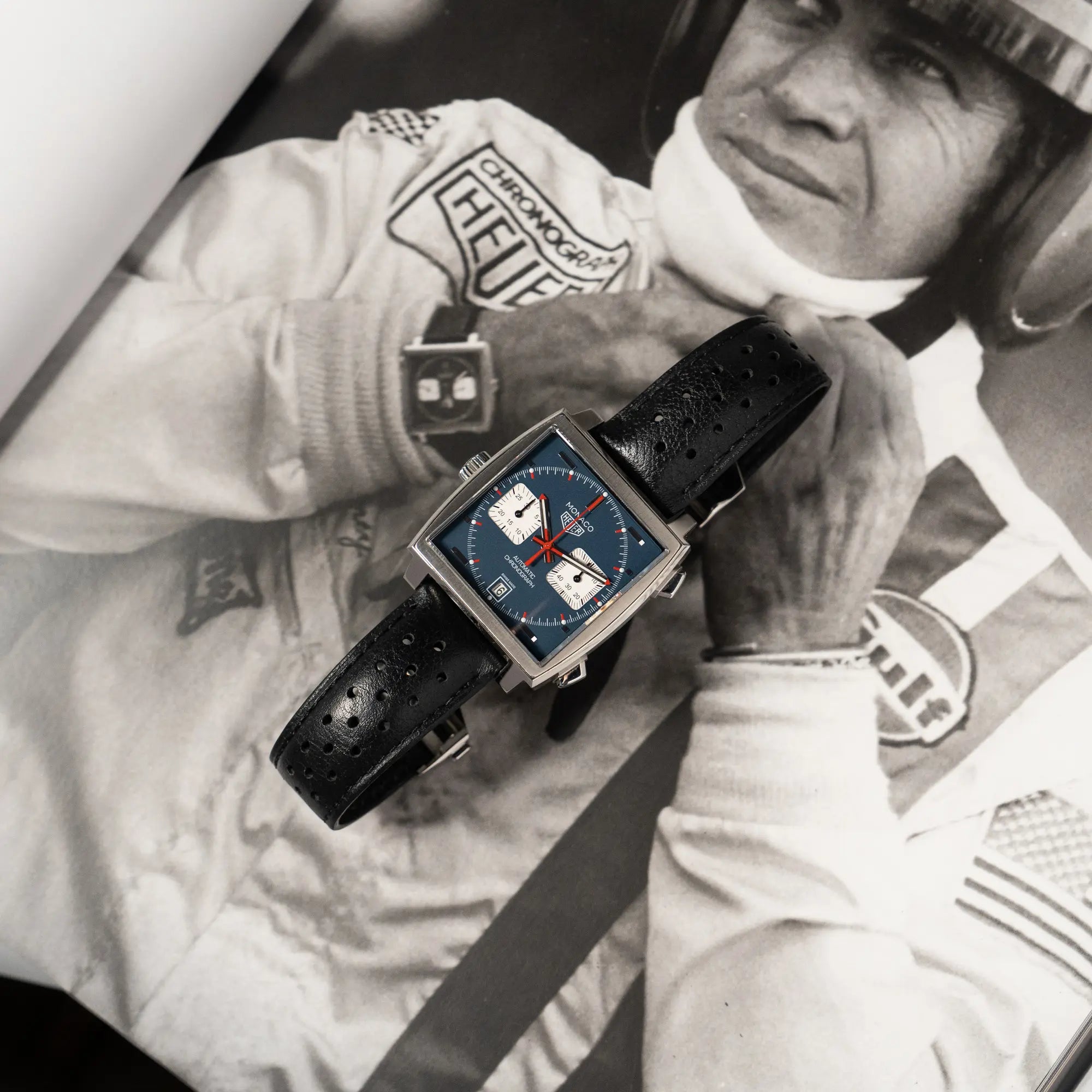 Produktfotografie des Tag Heuer Monaco Calibre 11 Chronographen CAW211P mit dem klassischen blauen Zifferblatt und roten Details auf Zeigern und Indizes mit schwarz-weißem Steve McQueen Foto als Hintergrund